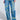 Someone is wearing JETSETTER EMBROIDERY WIDE LEG JEANS featuring bold yellow city names like "LONDON," "MILAN," and "TOKYO." They have a relaxed fit with knee rips. Paired with a colorful long-sleeved top and white shoes, these jeans offer a chic, urban vibe.
