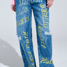 Someone is wearing JETSETTER EMBROIDERY WIDE LEG JEANS featuring bold yellow city names like "LONDON," "MILAN," and "TOKYO." They have a relaxed fit with knee rips. Paired with a colorful long-sleeved top and white shoes, these jeans offer a chic, urban vibe.