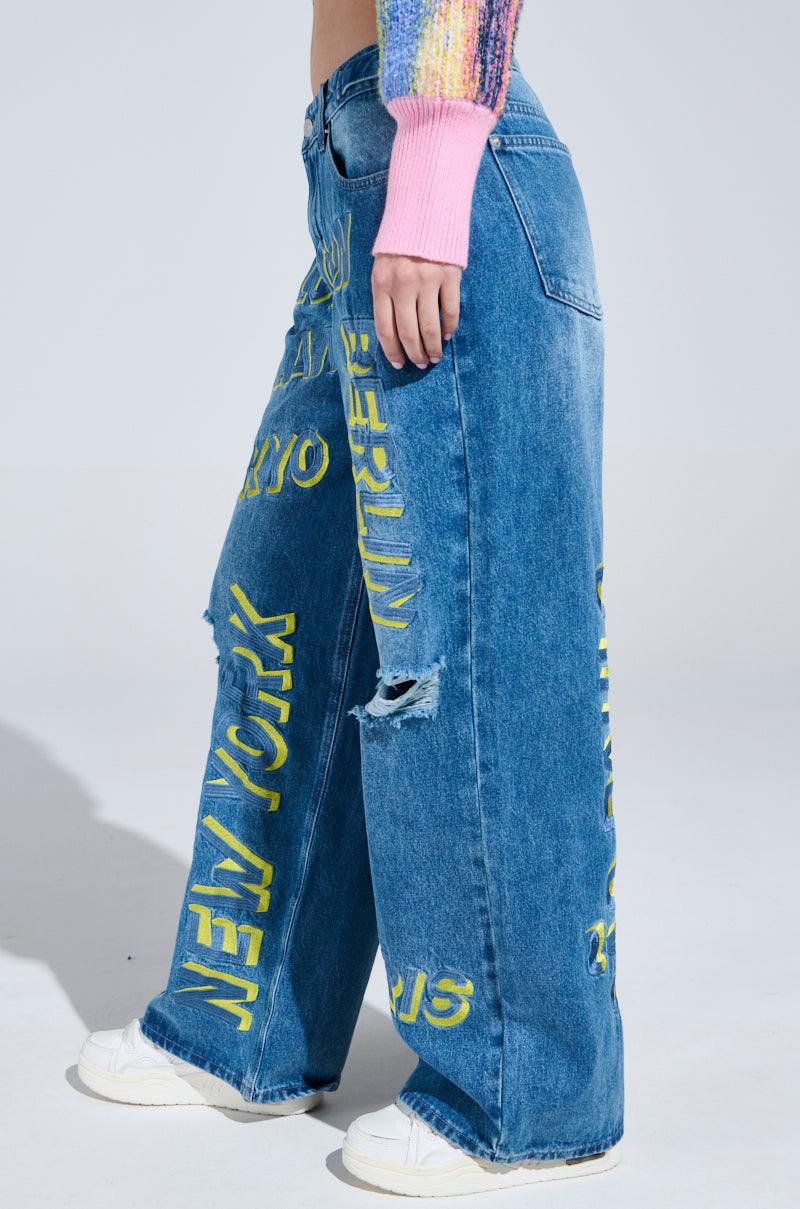Wearing JETSETTER EMBROIDERY WIDE LEG JEANS in blue, featuring wide legs and yellow city names like Berlin and New York with distressed details, this person completes their travel look with a colorful knit sweater and white sneakers.