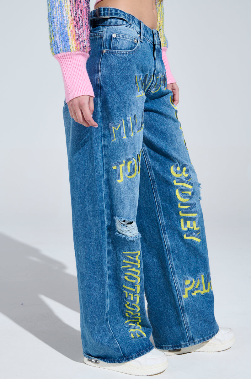 A person wearing Jetsetter Embroidery Wide Leg Jeans, adorned with vibrant city names like Milan, Tokyo, and Paris. These distressed baggy jeans pair perfectly with white sneakers and a colorful sweater with pink cuffs for an ideal travel style ensemble.