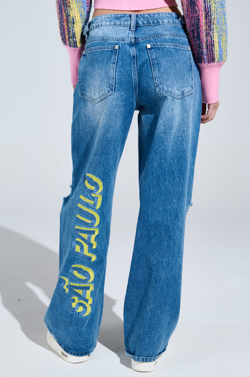 Rear view of a person sporting Jetsetter Embroidery Wide Leg Jeans with "São Paulo" in yellow on the right leg, paired with a colorful, textured sweater and white shoes, exuding jetsetter vibes.