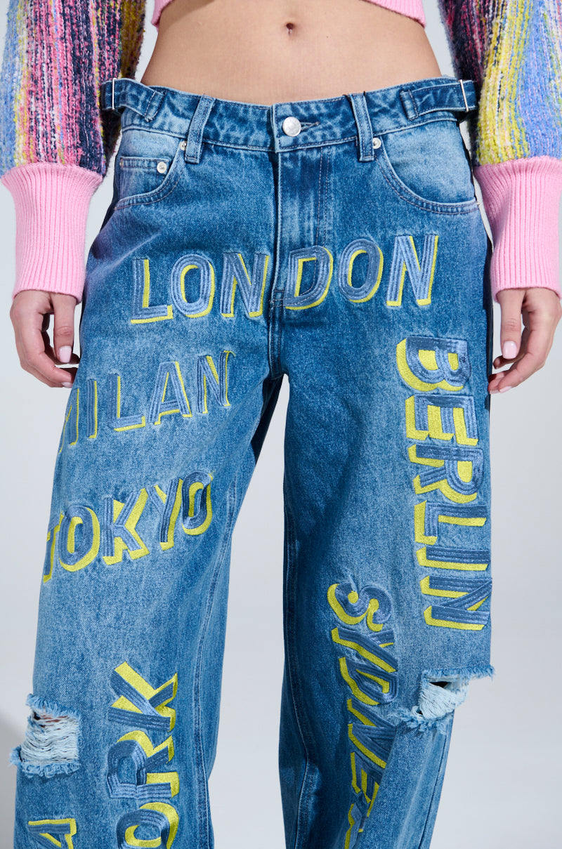 The person is flaunting Jetsetter Embroidery Wide Leg Jeans, showcasing yellow city names such as London, Milan, Tokyo, and Berlin with chic rips above the knee. Paired flawlessly with a vibrant sweater featuring pink sleeves, this ensemble embodies style and sophistication.