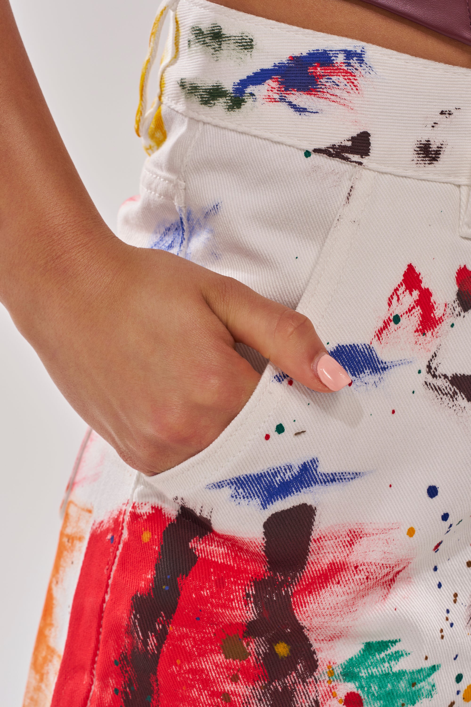 A hand with pink nail polish rests in the pocket of white PAINTED ON RELAXED DENIM PANT, splattered with colorful paint in red, blue, green, and black.