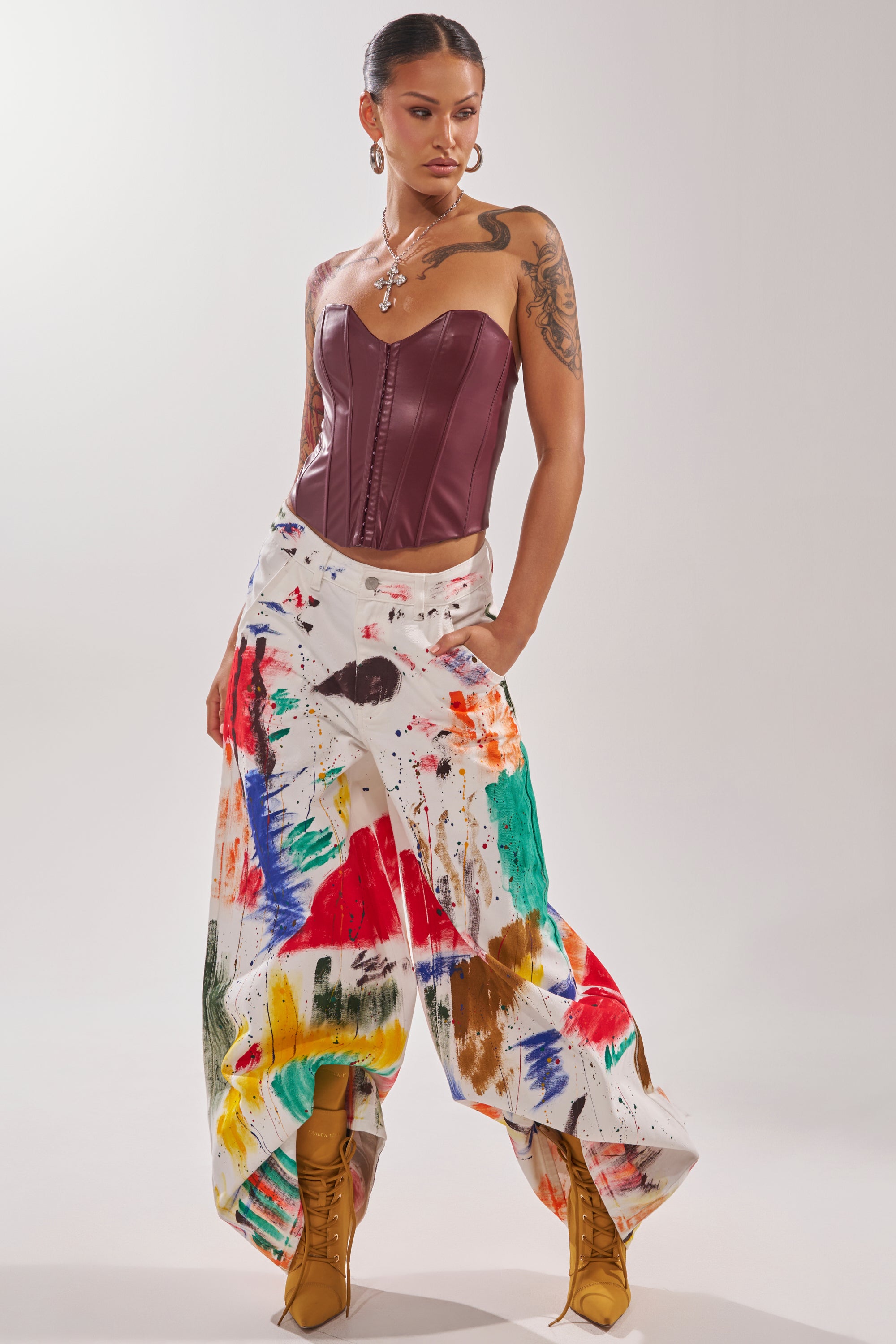 A woman in a burgundy corset top, PAINTED ON RELAXED DENIM PANT with colorful splatters, tan lace-up boots, large hoop earrings, and layered necklaces stands confidently against a plain background.