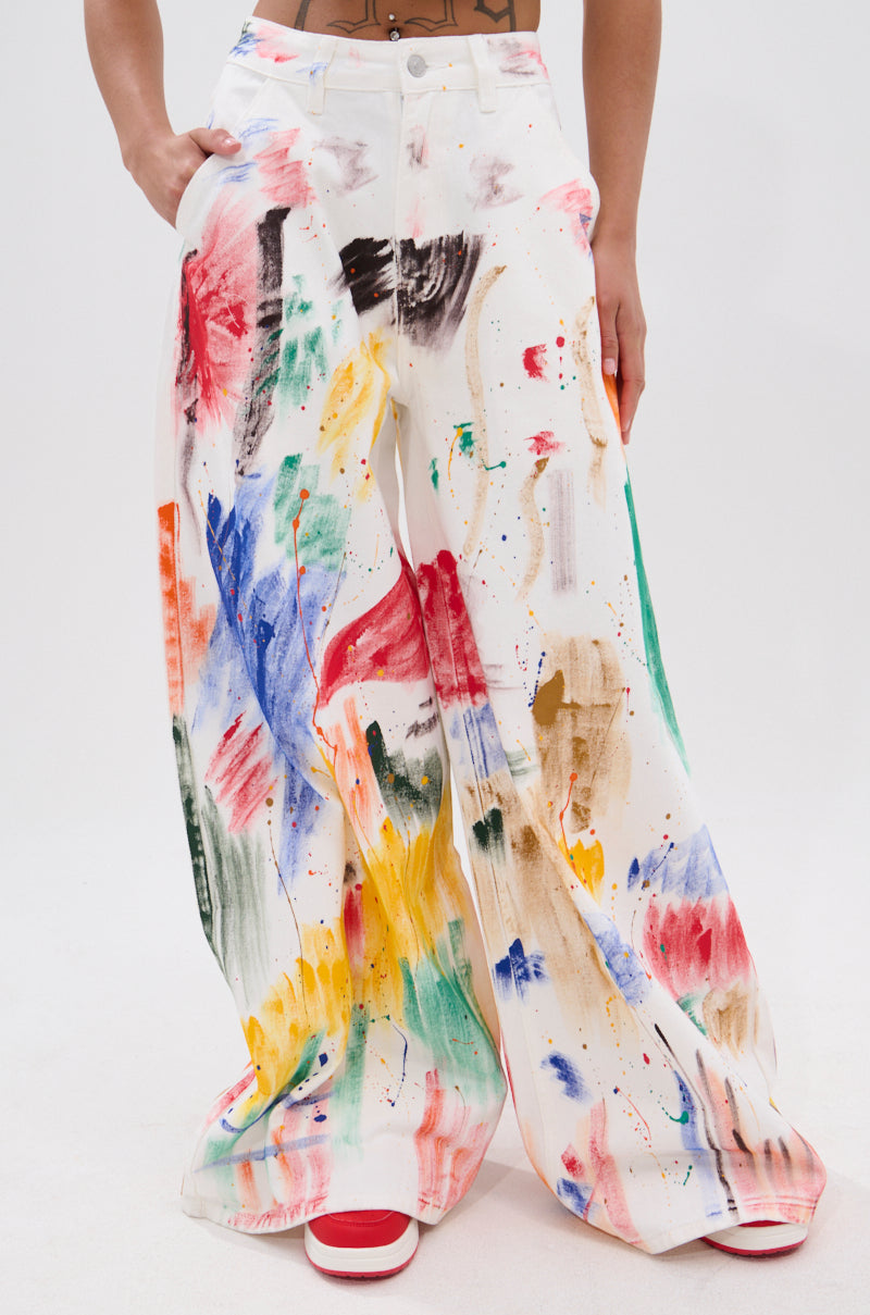 Model wears PAINTED ON RELAXED DENIM PANT—white, high-rise, wide-leg denim with painterly brush stroke splatters in red, blue, yellow, green, and brown—paired with red sneakers against a plain white background.