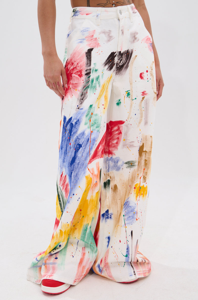 Wearing the PAINTED ON RELAXED DENIM PANT, featuring a high-rise fit and wide legs with painterly brush stroke designs in white, paired with red and white sneakers. The plain white background highlights the look.