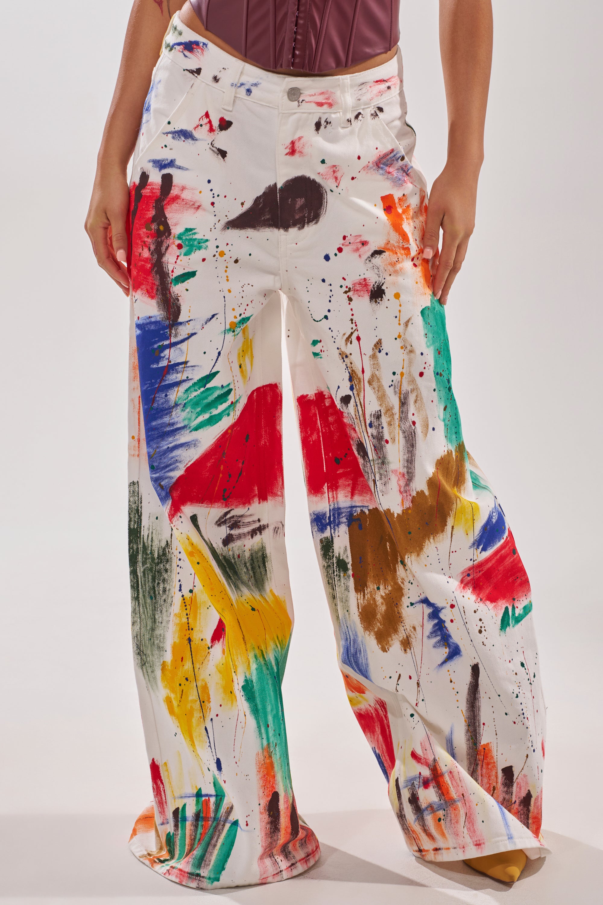 A person models the PAINTED ON RELAXED DENIM PANT, featuring loose-fitting white denim with a wide-leg silhouette and colorful paint splatters, standing against a plain background with hands on their hips.