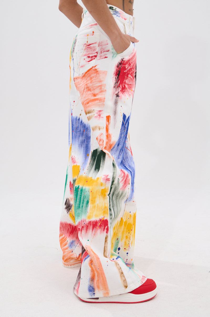 A person stands sideways in high-rise, wide-leg PAINTED ON RELAXED DENIM PANT featuring colorful brush stroke splatters, paired with red-and-white sneakers. Their hand is in their pocket against a plain white background.