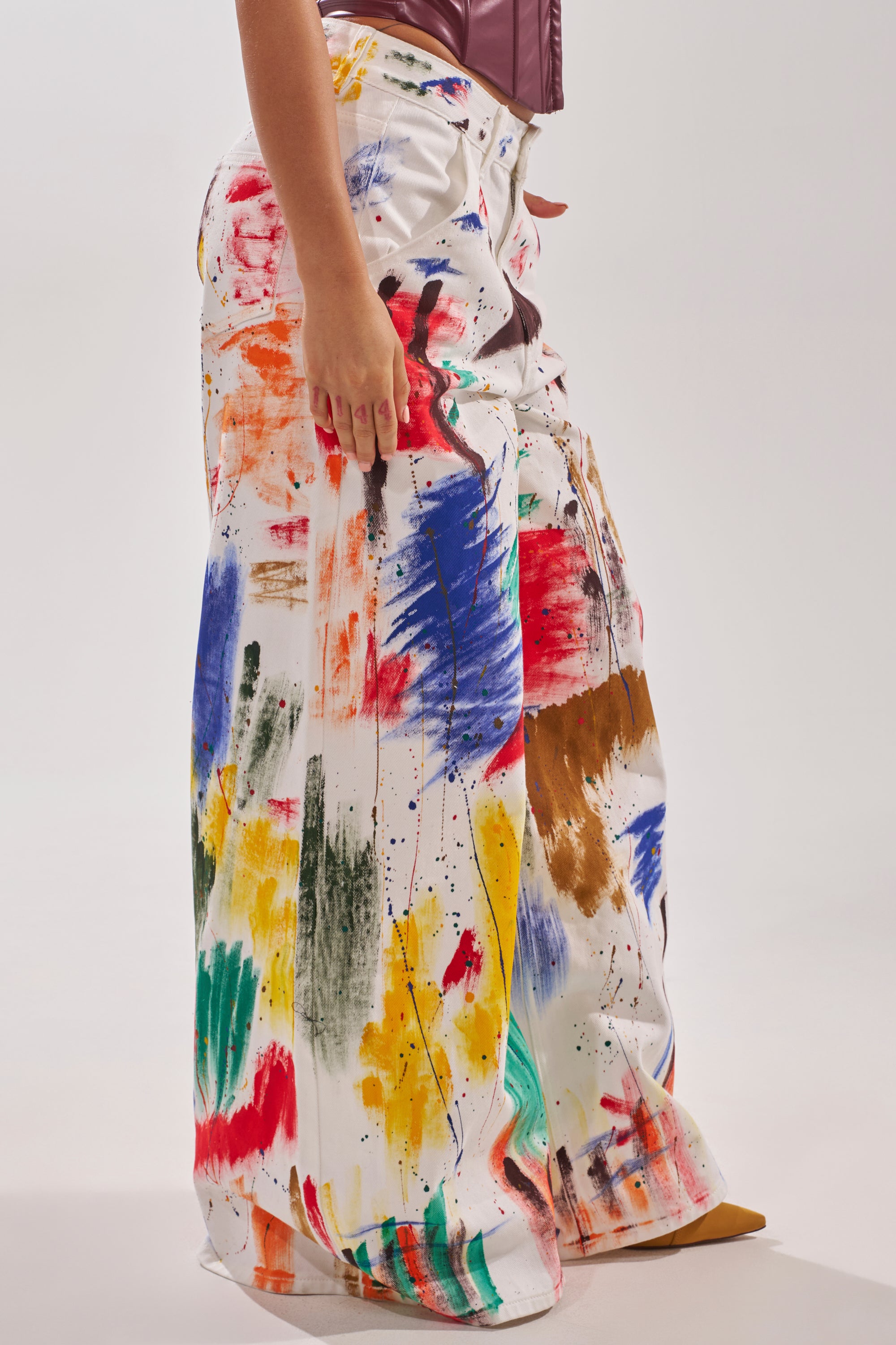A person models the PAINTED ON RELAXED DENIM PANT, wide-leg jeans with colorful abstract paint splatters and brushstrokes in red, blue, yellow, green, orange, and brown against a plain light background.