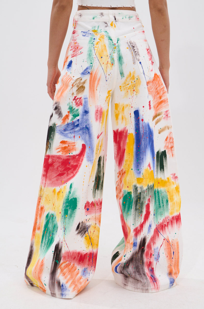 A person wearing the PAINTED ON RELAXED DENIM PANT, a high-rise, wide-leg white pant with a bold, colorful painterly brush stroke design, shown from the back against a plain white background.
