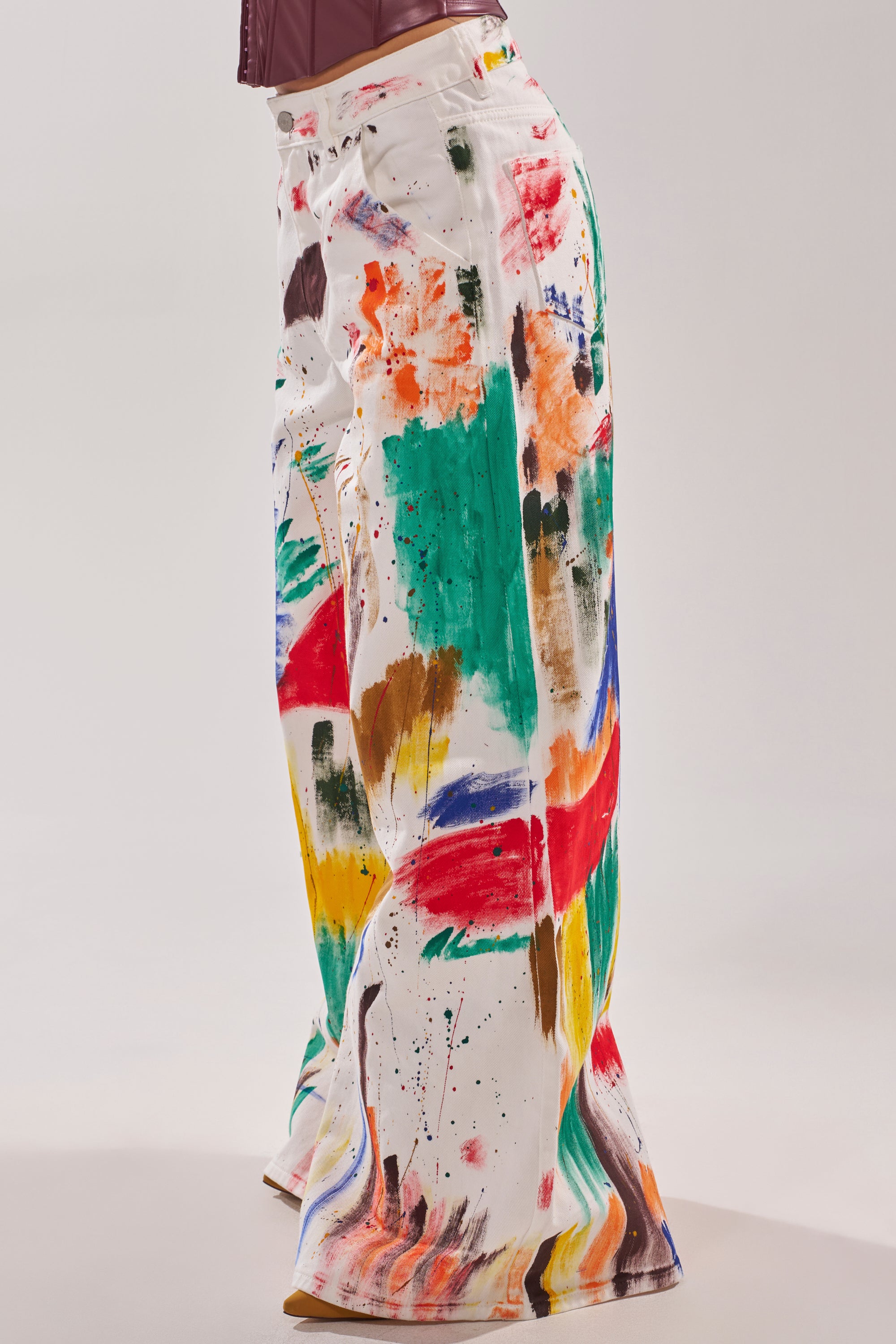 Someone models the PAINTED ON RELAXED DENIM PANT, featuring wide-leg jeans with colorful abstract splatters and brushstrokes, set against a plain light background for an artistic style statement.