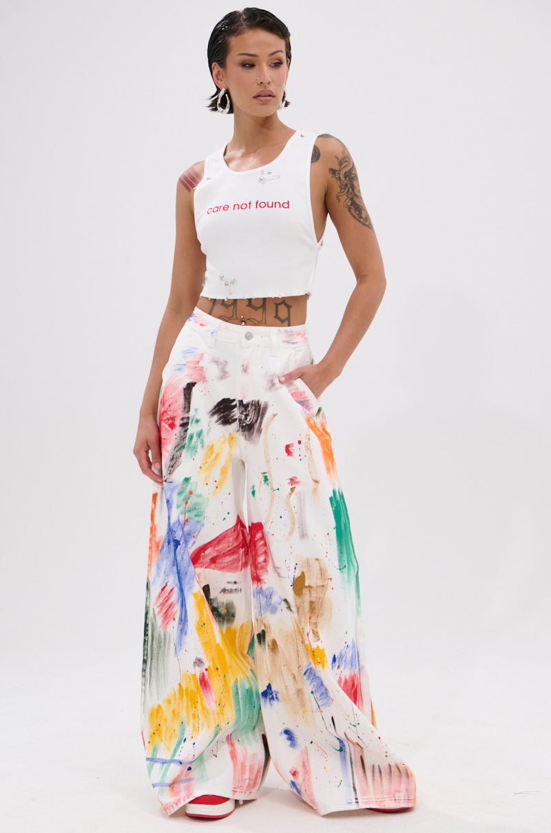 A woman with short slicked-back hair and arm tattoos stands against a white background wearing a "care not found" white crop top and PAINTED ON RELAXED DENIM PANT featuring colorful brushstroke splatters.