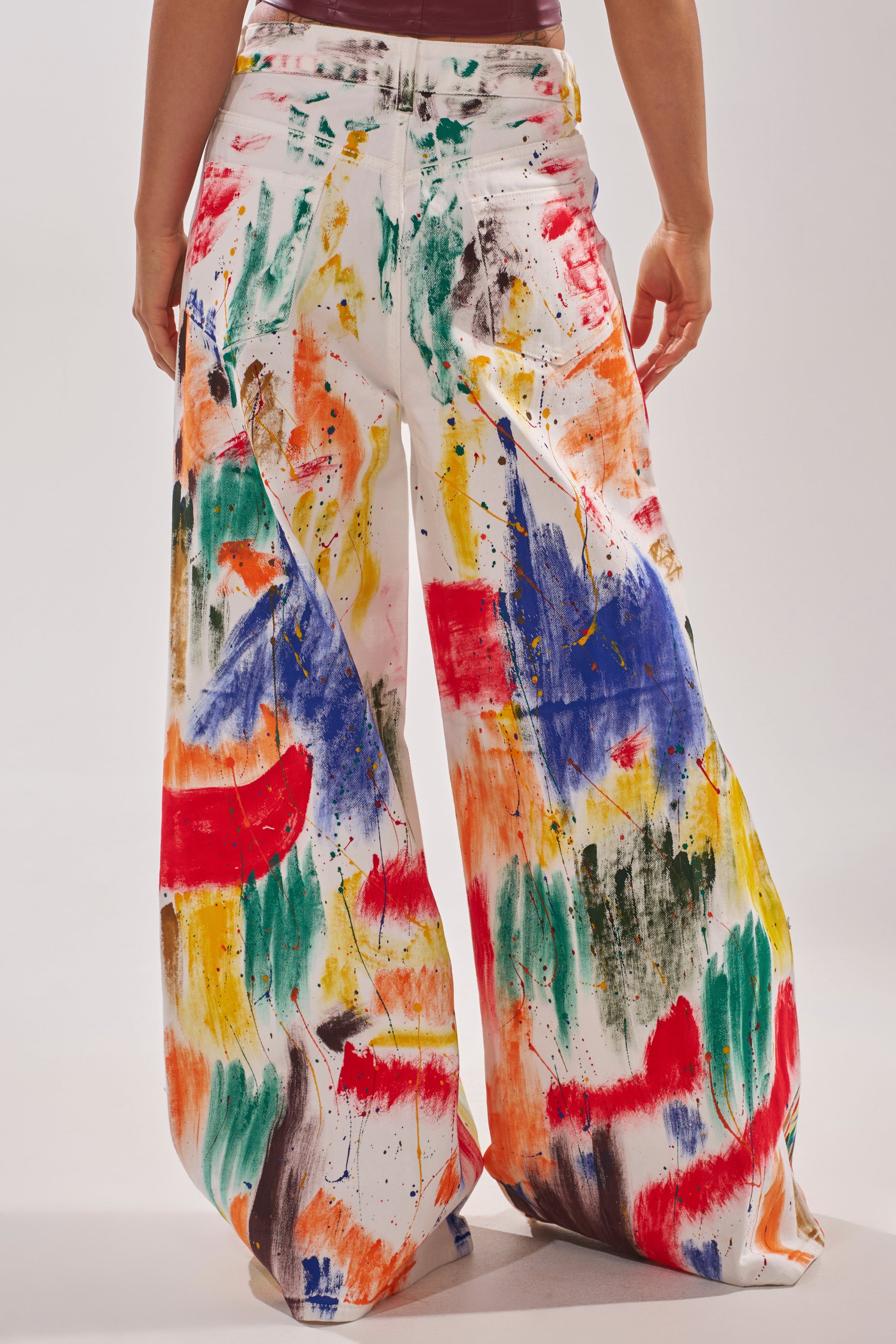 A person models the PAINTED ON RELAXED DENIM PANT, featuring wide-leg jeans with a vibrant, abstract paint splatter design in red, blue, green, yellow, and black against a plain background.