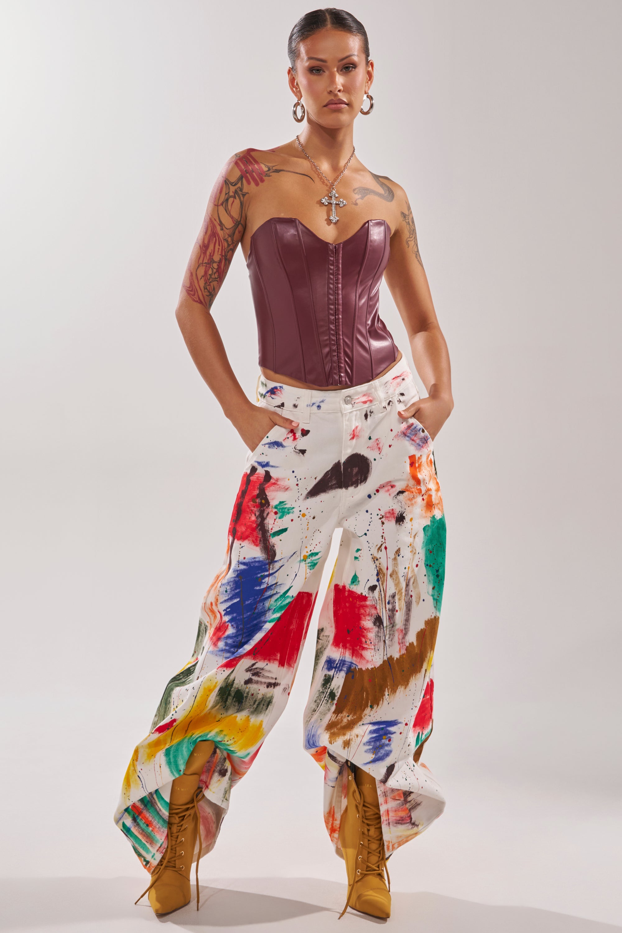 A person stands confidently in PAINTED ON RELAXED DENIM PANT with colorful splatters, a maroon leather corset, tan lace-up boots, hoop earrings, a large cross necklace, and visible arm tattoos.