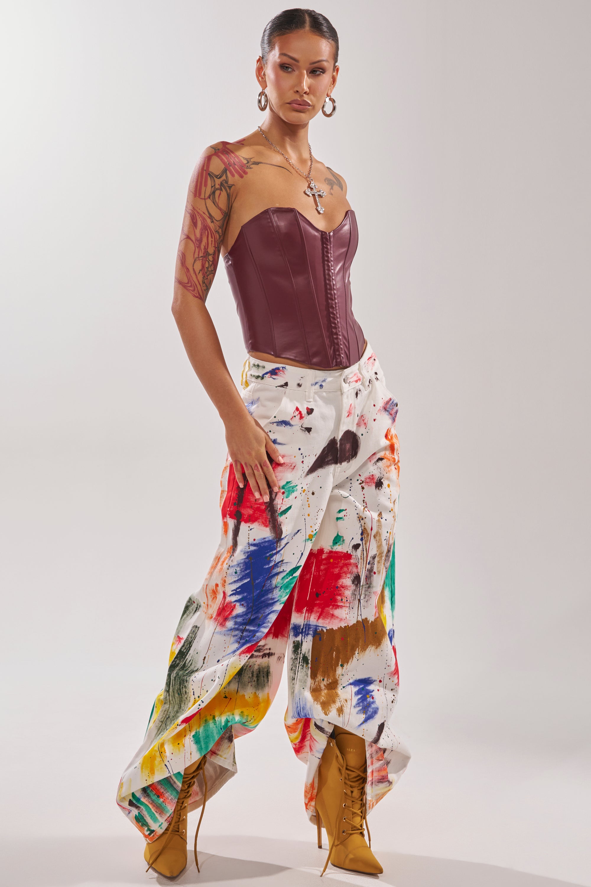 A woman exudes confidence in PAINTED ON RELAXED DENIM PANT splattered with colors, paired with a strapless burgundy corset, tan lace-up boots, hoop earrings, layered necklaces, her tattooed arm visible and hair styled sleekly.