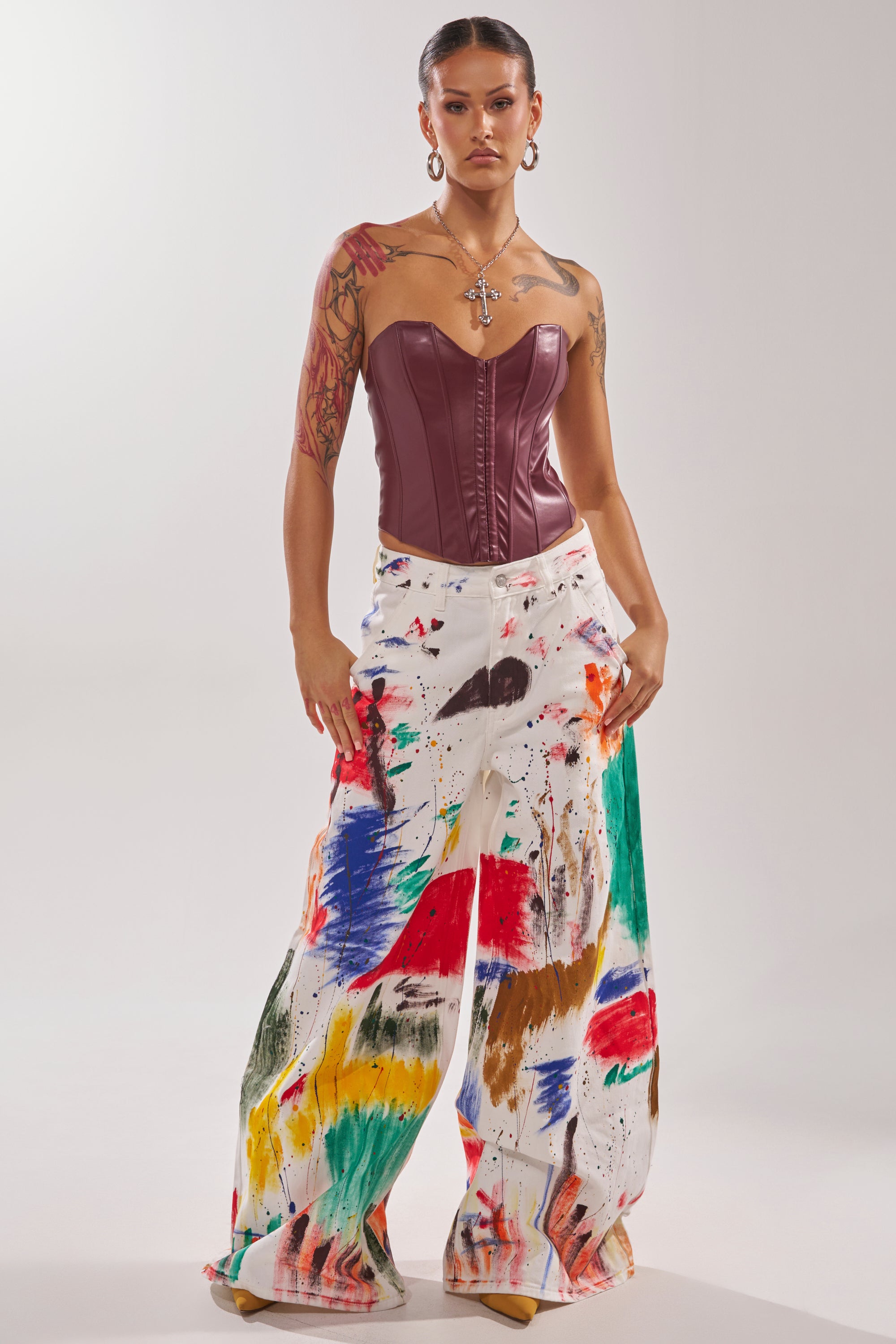A woman stands confidently in the PAINTED ON RELAXED DENIM PANT paired with a strapless maroon corset. She wears hoop earrings, a cross necklace, and displays arm tattoos, set against a plain studio background.