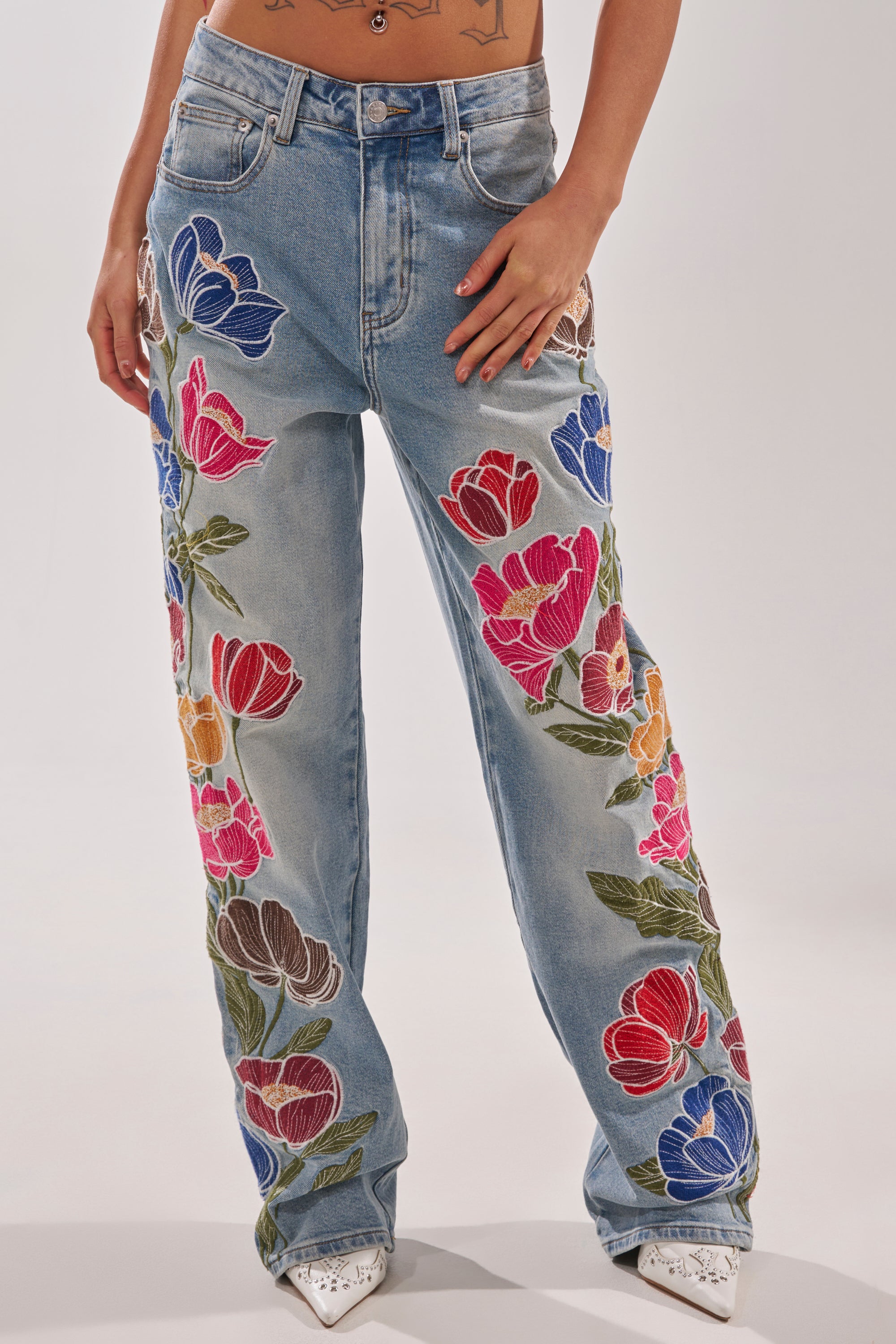 A person wears INTO THE GARDEN FLORAL EMBROIDERED DENIM JEANS—high-rise, straight-leg jeans with floral embroidery—styled with white pointed-toe shoes against a plain background. 0