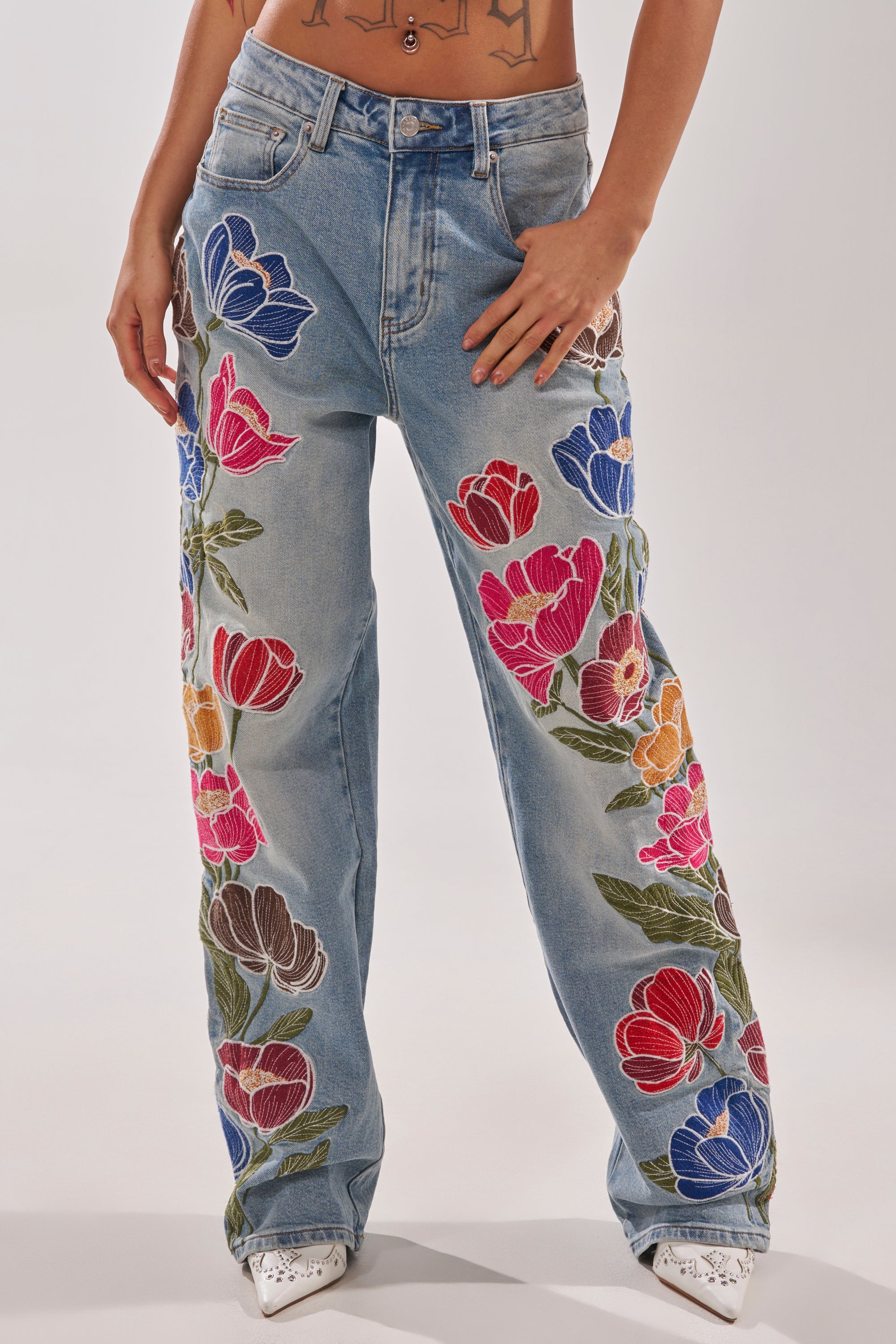 A person wears INTO THE GARDEN FLORAL EMBROIDERED DENIM JEANS in light blue, featuring vibrant floral embroidery and a high-rise wide-leg fit, paired with white pointed-toe shoes. Only the torso and legs are visible. 1