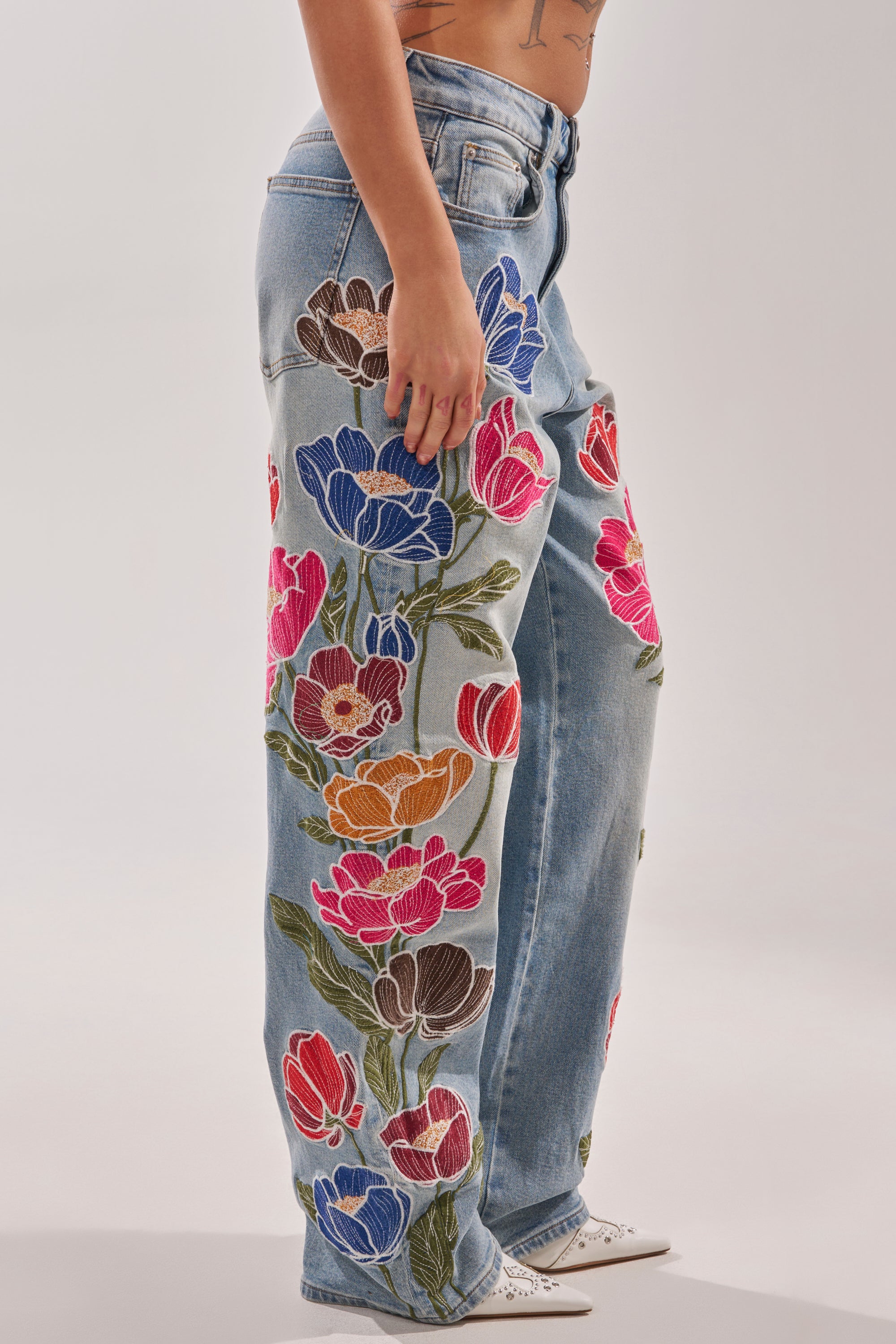 Wearing INTO THE GARDEN FLORAL EMBROIDERED DENIM JEANS in light blue with bold red, pink, blue, orange, and brown flower embroidery, the person stands sideways showing tattooed arms and white shoes. 2