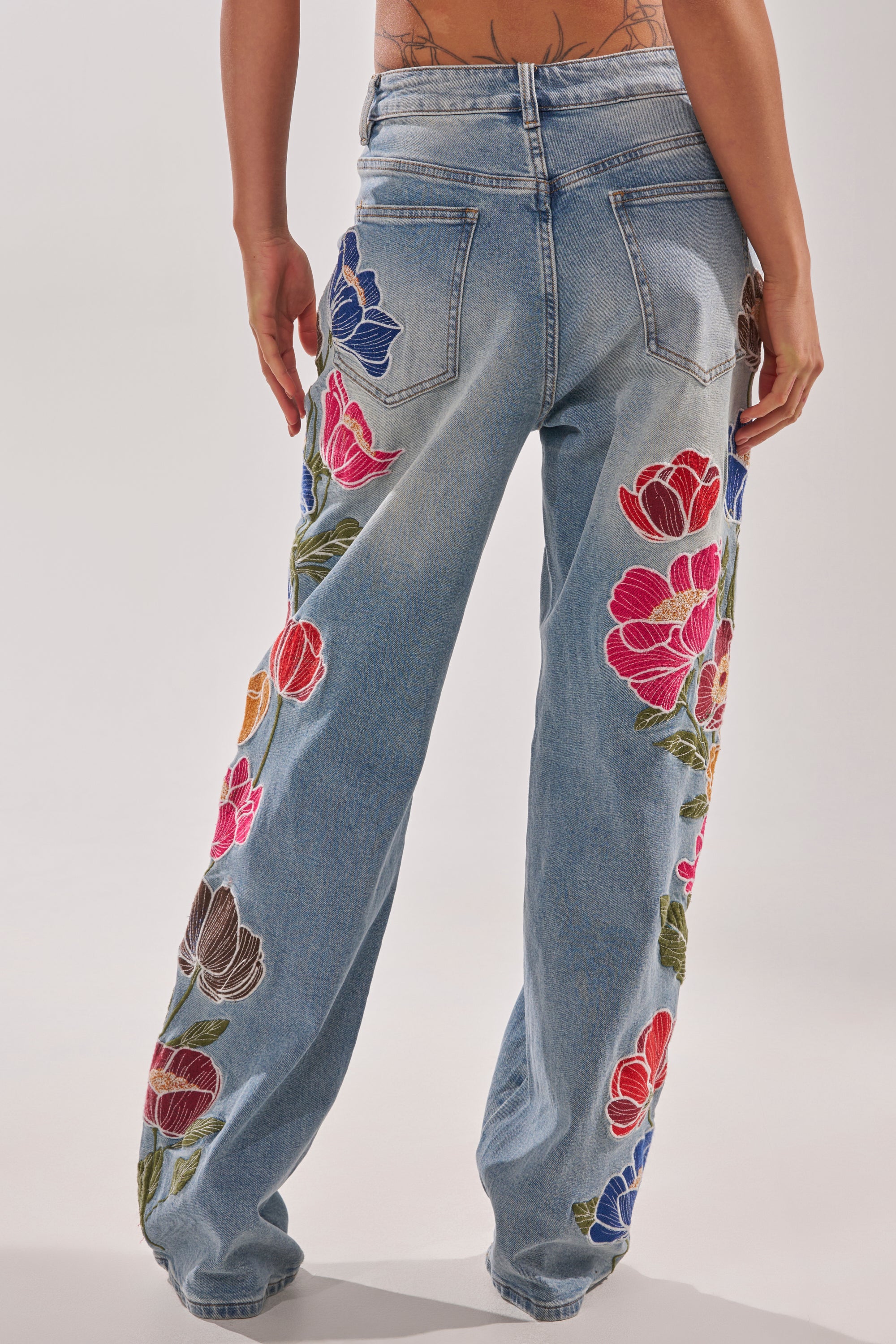 A person models the INTO THE GARDEN FLORAL EMBROIDERED DENIM JEANS, featuring boho-inspired, high-rise denim with colorful floral embroidery on the sides and back, shown from behind against a plain background. 3