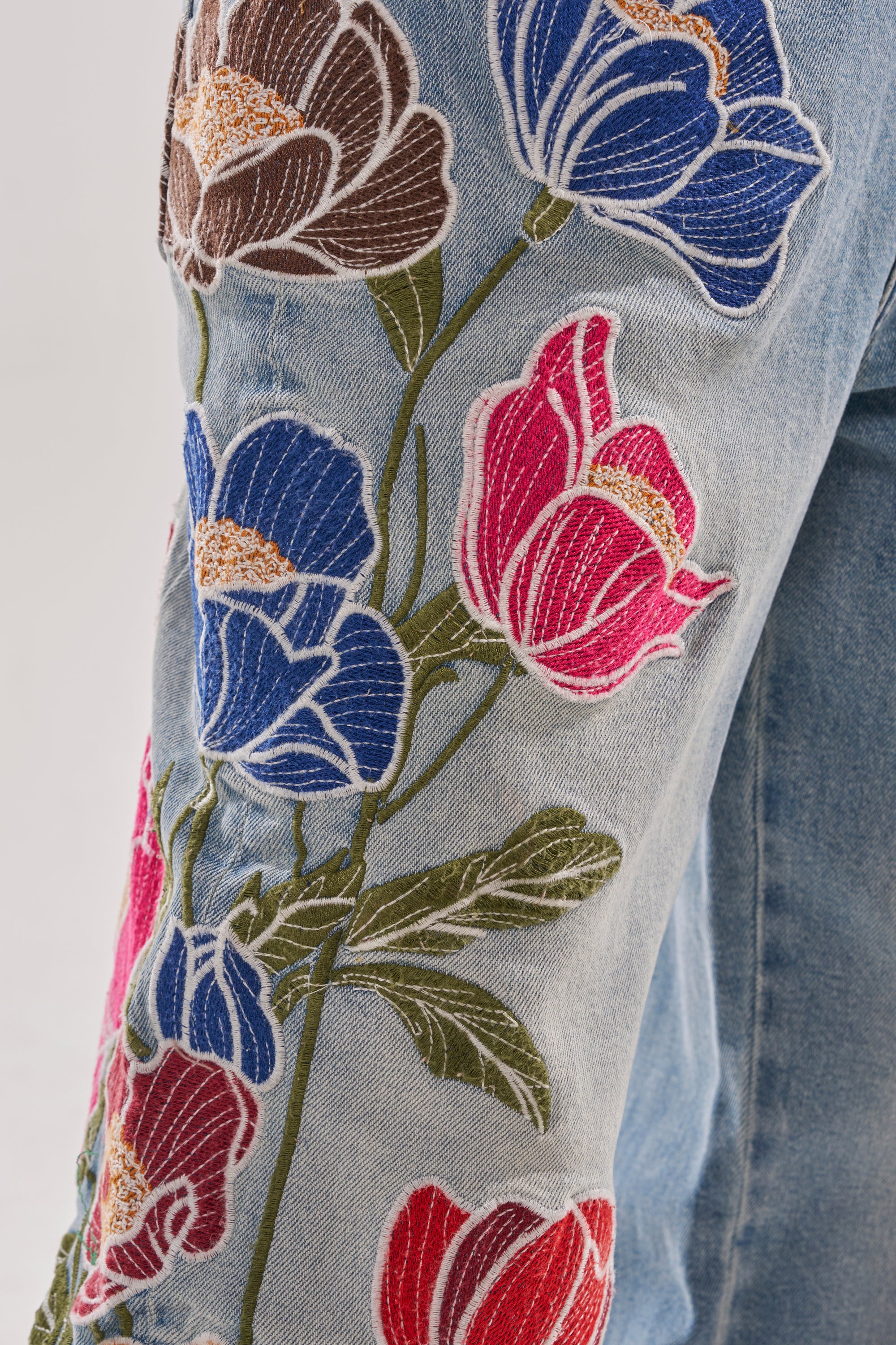 Close-up of INTO THE GARDEN FLORAL EMBROIDERED DENIM JEANS with red, pink, blue, and brown flowers plus green stems and leaves. These vibrant jeans feature a bold boho-inspired floral design across much of the denim. 7