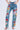 Someone pairs the INTO THE GARDEN FLORAL EMBROIDERED DENIM JEANS, featuring a high-rise light-wash design with colorful floral embroidery, with a white top. Hands rest casually in pockets, completing the look with black heeled sandals against a plain white backdrop.