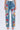 A person is wearing the INTO THE GARDEN FLORAL EMBROIDERED DENIM JEANS with vibrant red, orange, pink, and blue floral designs. The light-wash denim is paired with a white top and open-toed heels against a plain white background.