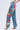 Someone wears the INTO THE GARDEN FLORAL EMBROIDERED DENIM JEANS with light-wash denim and colorful side embroidery featuring red, blue, yellow, and pink flowers. These high-rise jeans are styled with open-toe, high-heeled shoes against a plain white backdrop.