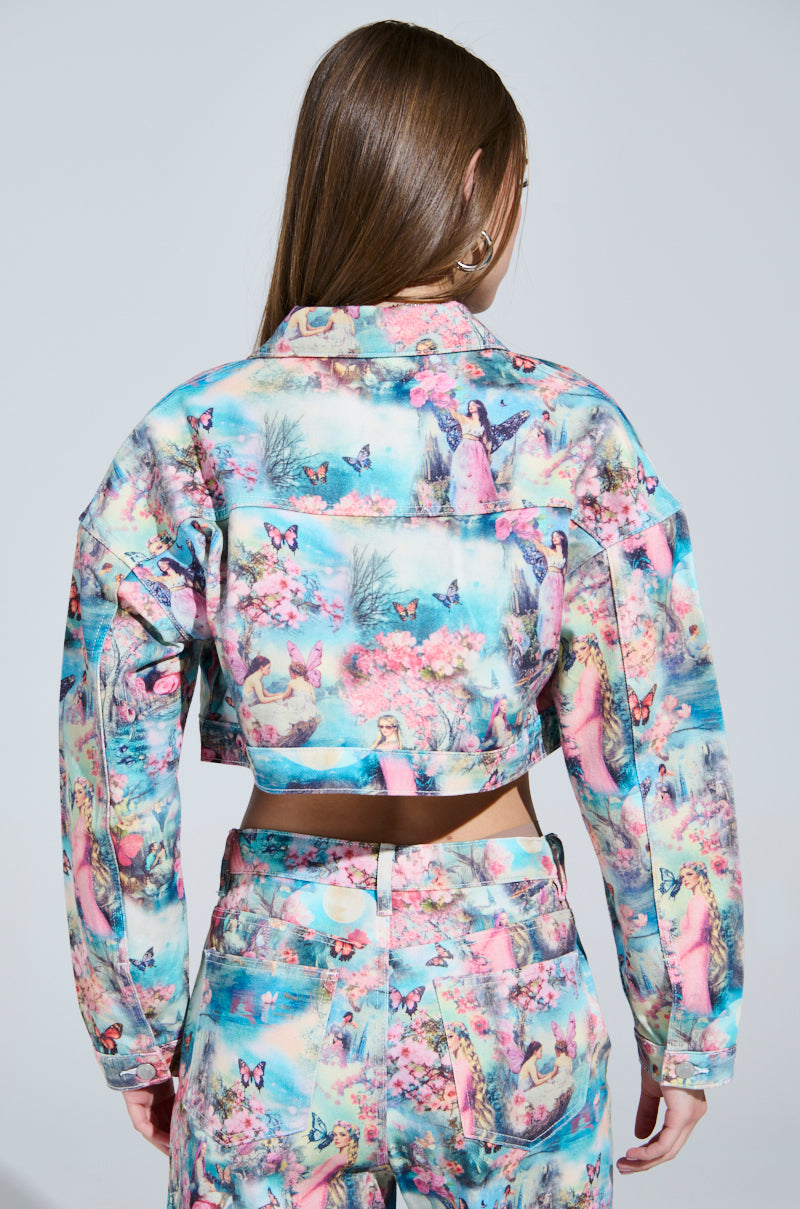 A person with long brown hair faces away, wearing the FEMALE FANTASY PRINTED DENIM JACKET and matching pants. The set features a cropped silhouette adorned with whimsical butterflies, flowers, and figures in a pastel candy-colored design.