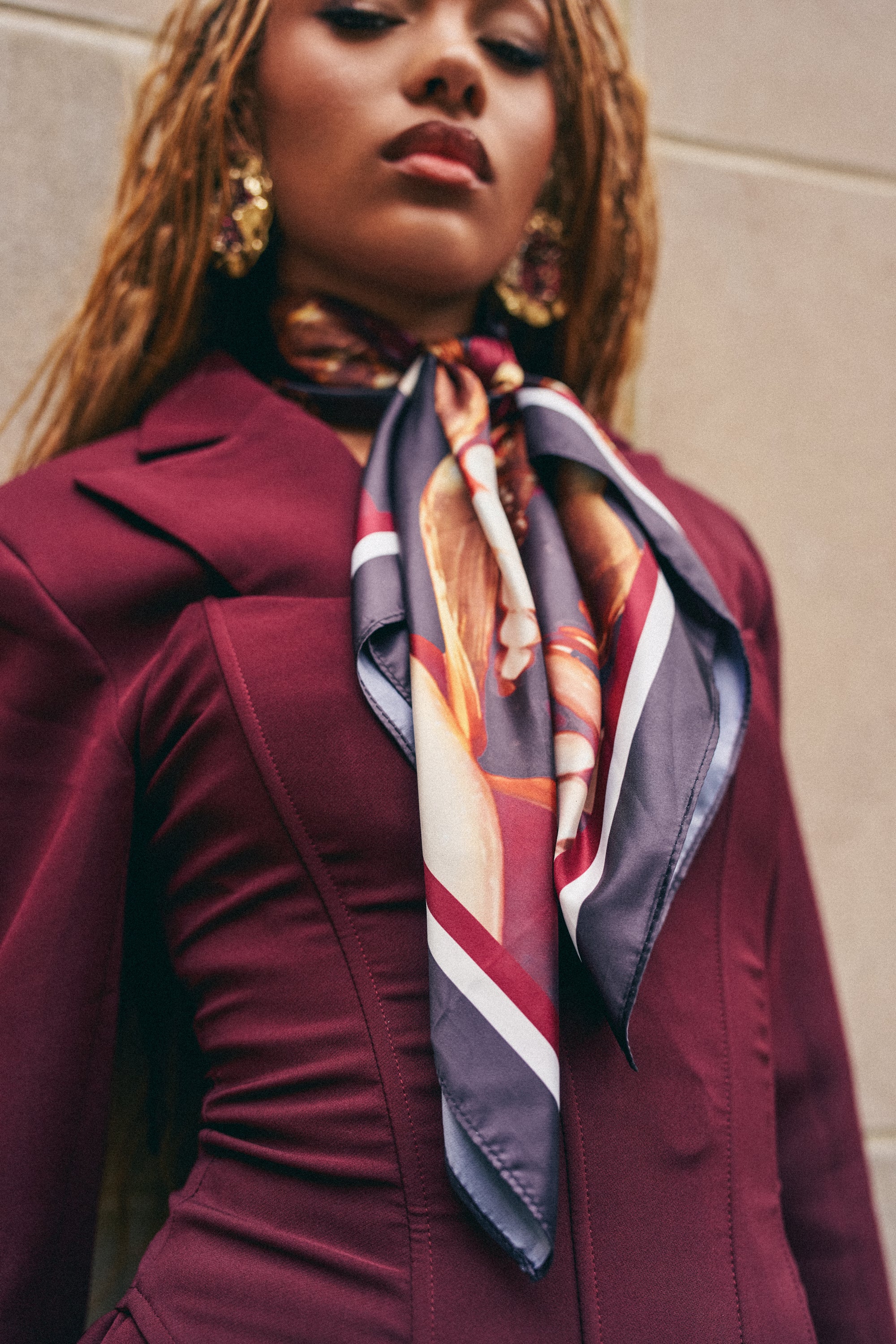A woman stands confidently against a beige wall, looking at the camera, wearing a burgundy blazer paired with the JUST SMILE SATIN SCARF featuring a bold lips and grills print. The photo is captured from a low angle to emphasize her outfit and accessories.