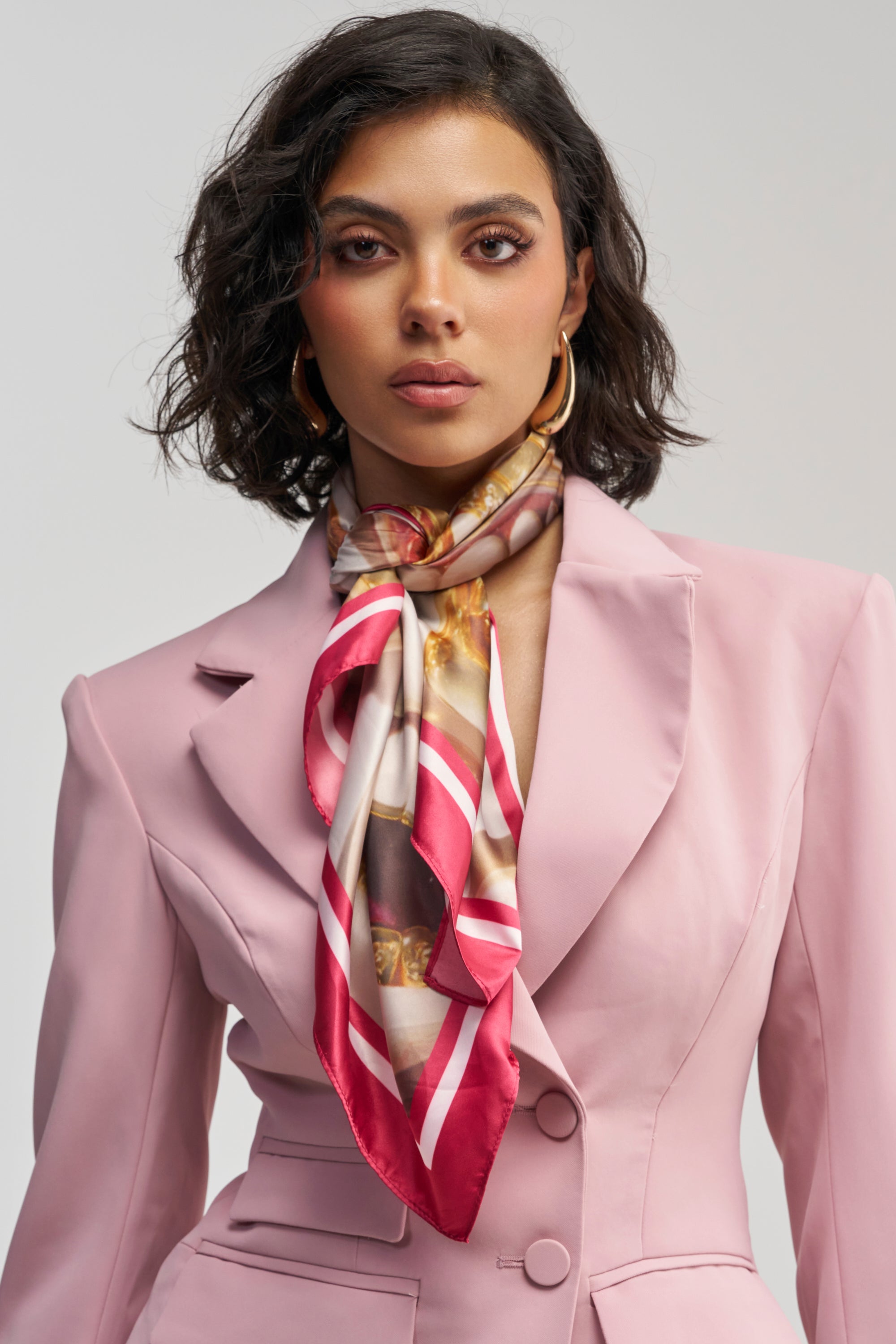 A woman with short dark hair wears a light pink blazer and the MOUTHY SATIN PRINTED SCARF, which features red, white, and gold patterns, standing against a plain background.