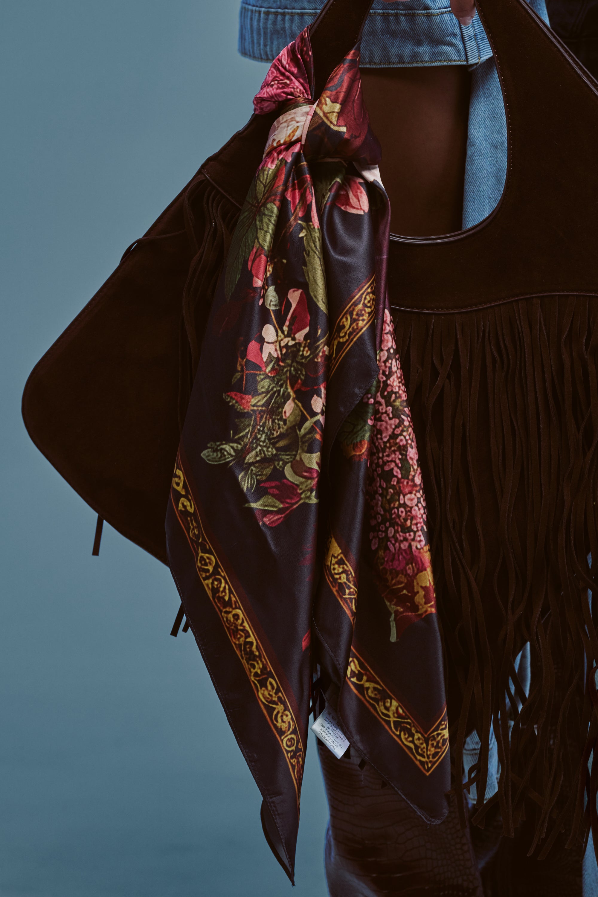 A black fringed bag features the DARK FLORAL SCARF—a vibrant black floral satin scarf—tied elegantly to its handle, displayed against a blue background. Touches of denim fabric peek through at the top of the image, making this chic accessory set ideal for any stylish summer outing.