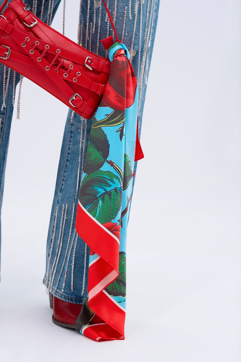 A person wears blue jeans with silver details, red shoes, and holds a red handbag accented by the ROSEY VIBE SATIN SCARF tied to it. The background is plain and light-colored.