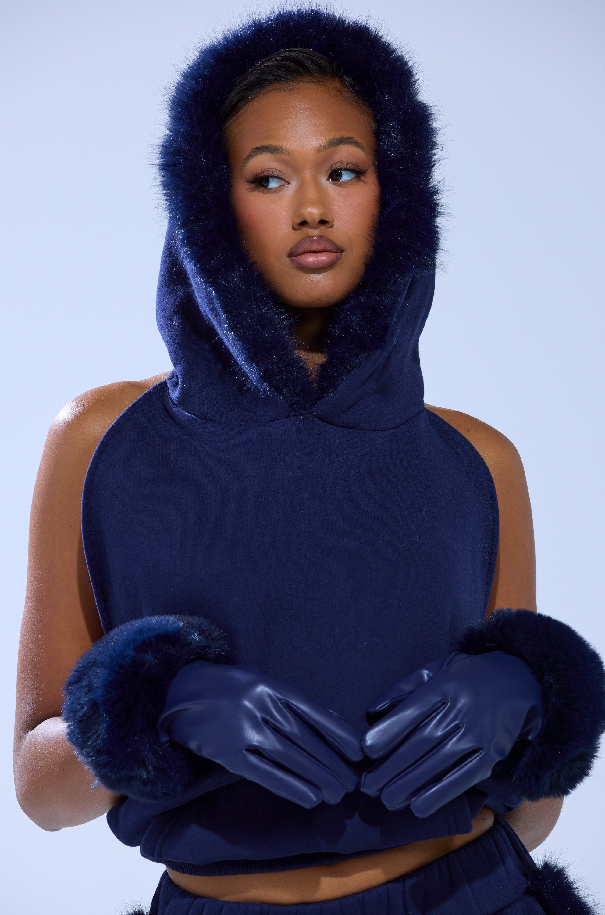 A woman wearing TOO CHIC FAUX FUR GLOVES—navy blue touchscreen gloves with fur cuffs—paired with a sleeveless navy blue hooded top, looking off to the side against a light blue background. 4