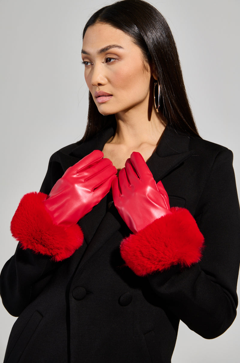 A person wearing a black coat with large red faux fur cuffs and the ALL I WANT IS MONEY FUR GLOVES stands against a plain background. They have long dark hair, are looking to the side, and don stylish large hoop earrings.