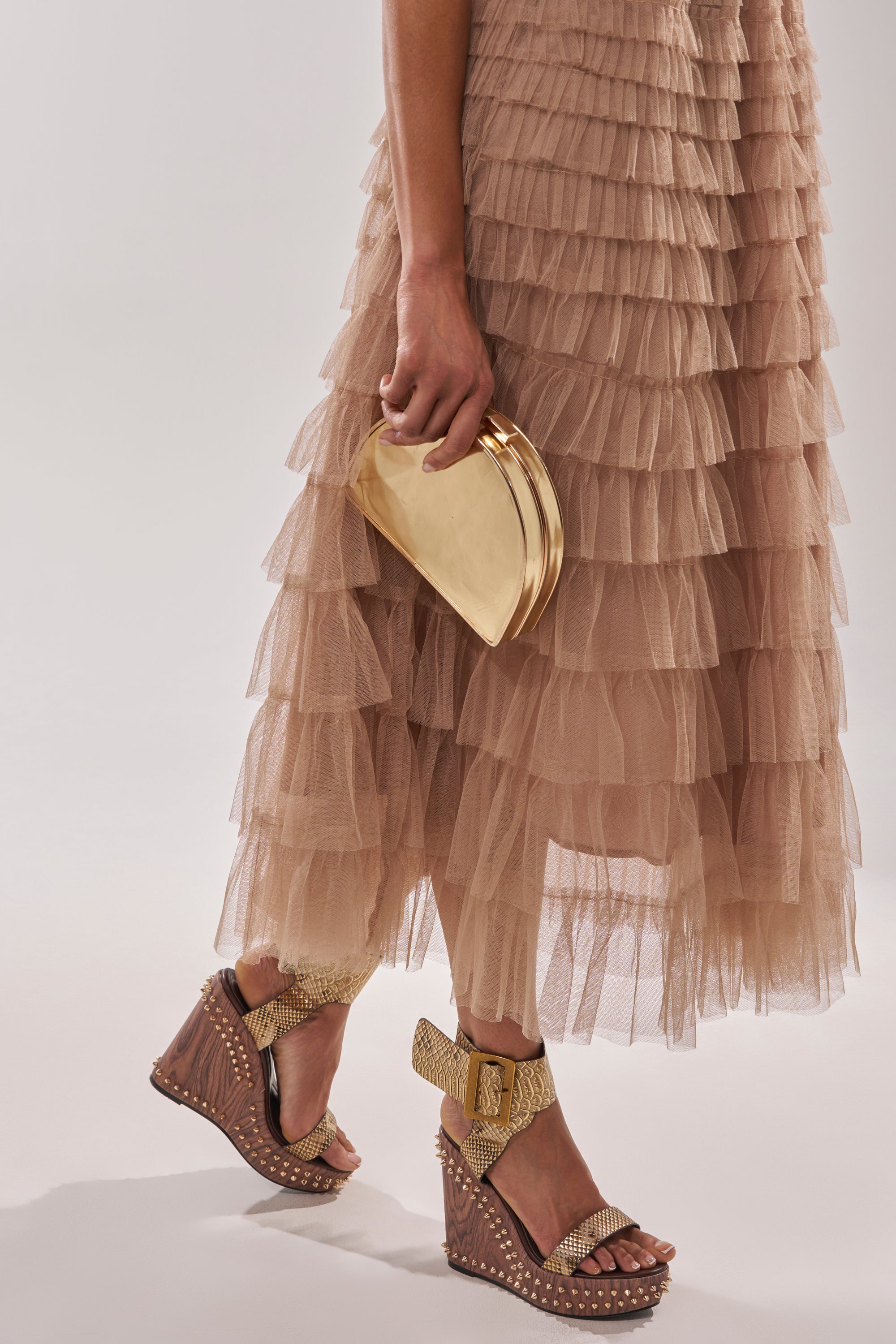 Wearing a layered neutral tulle skirt and holding a gold clutch, the person pairs their look with AKIRA GOLD SLOANE BROWN STUD AND SNAKE WEDGE SANDALS featuring bows and studded heels against a plain, light background. 1