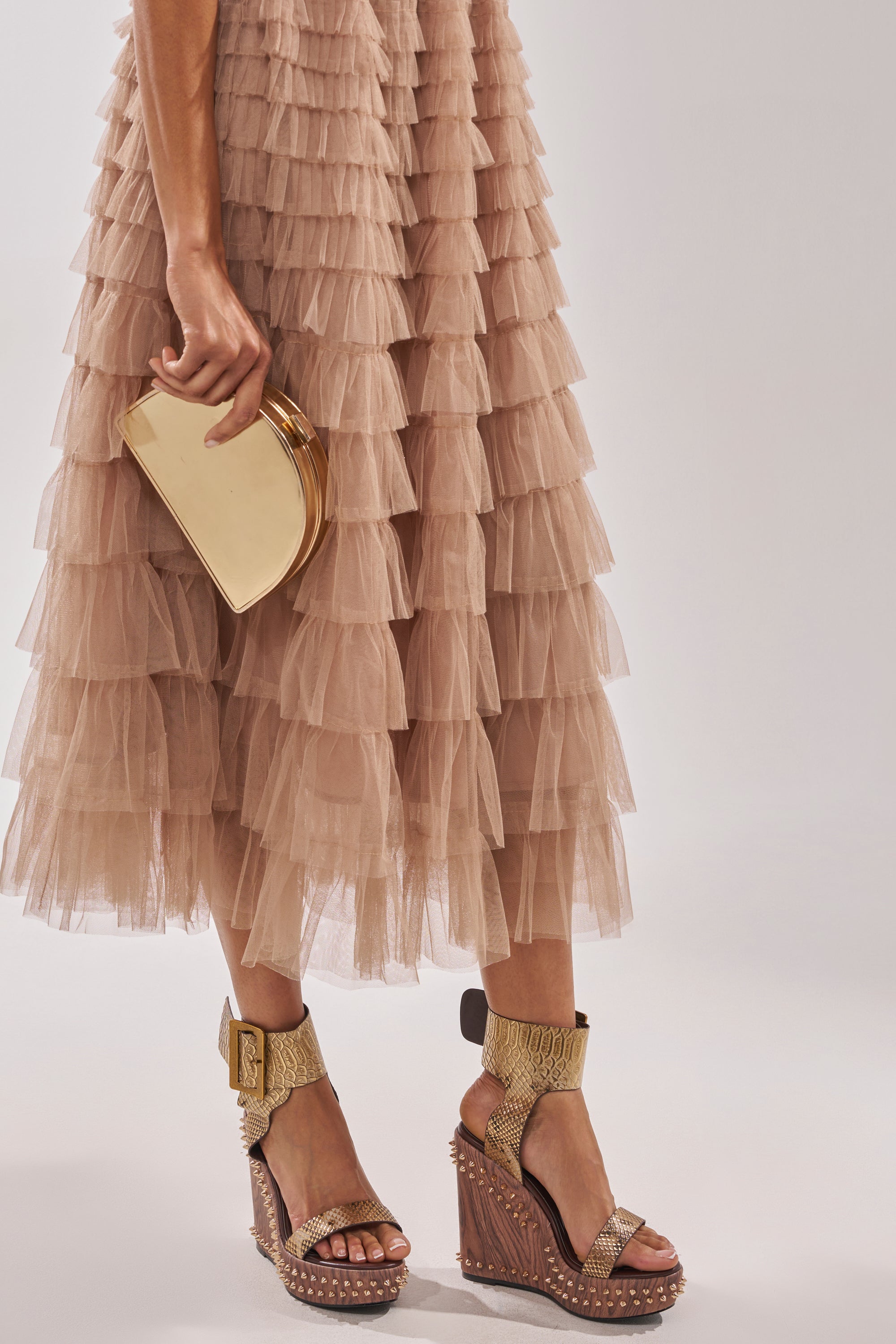 A woman, shown from the lower legs down, holds a small gold clutch and wears the AKIRA GOLD SLOANE BROWN STUD AND SNAKE WEDGE SANDAL with metallic snakeskin straps and gold stud accents, paired with a tiered beige tulle skirt. 2