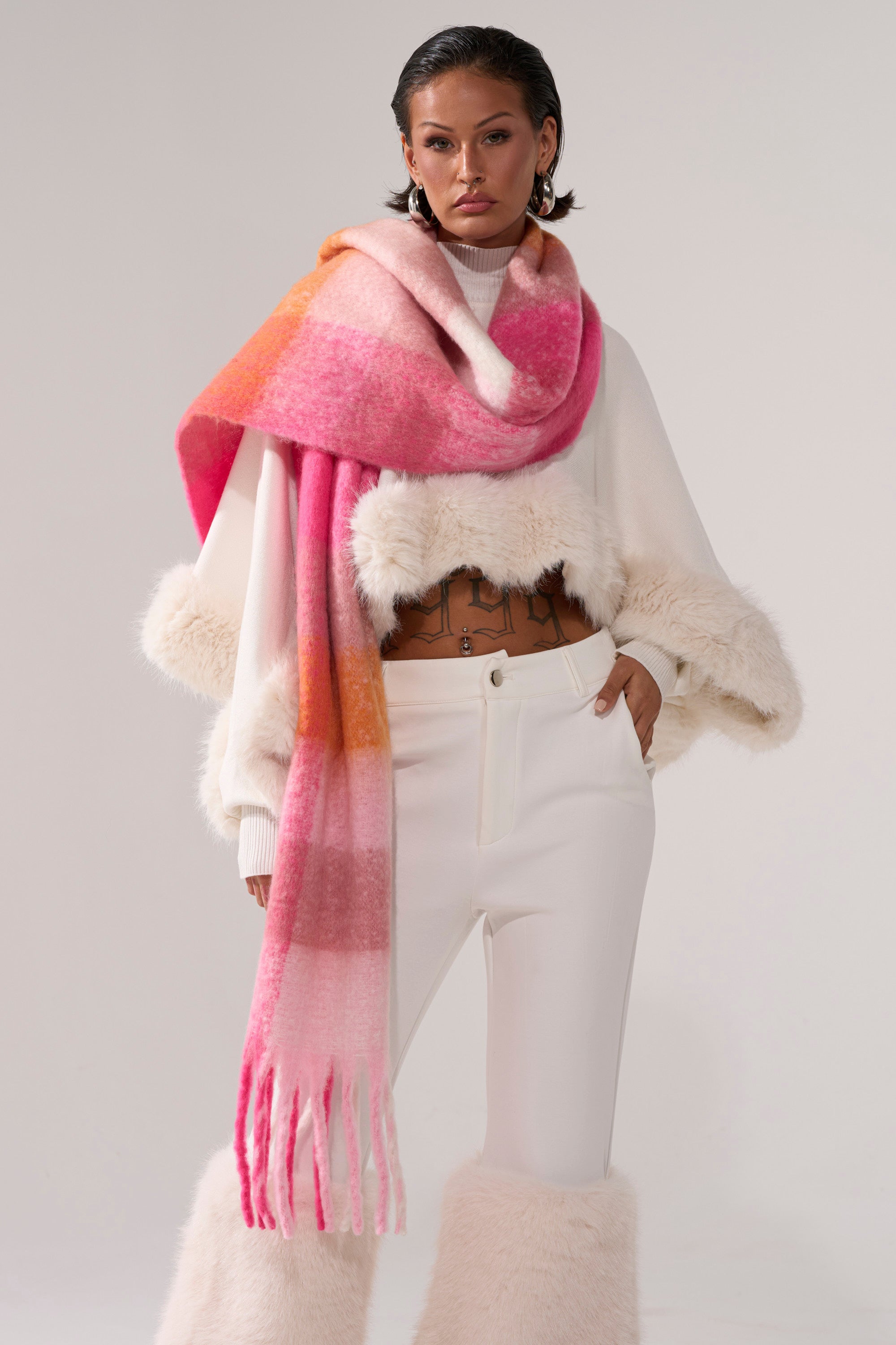 A woman with short dark hair poses confidently against a plain background, wearing a white furry sweater, white fur-cuff pants, and the IN MILAN BLANKET SCARF in a bold pink and orange ombre.