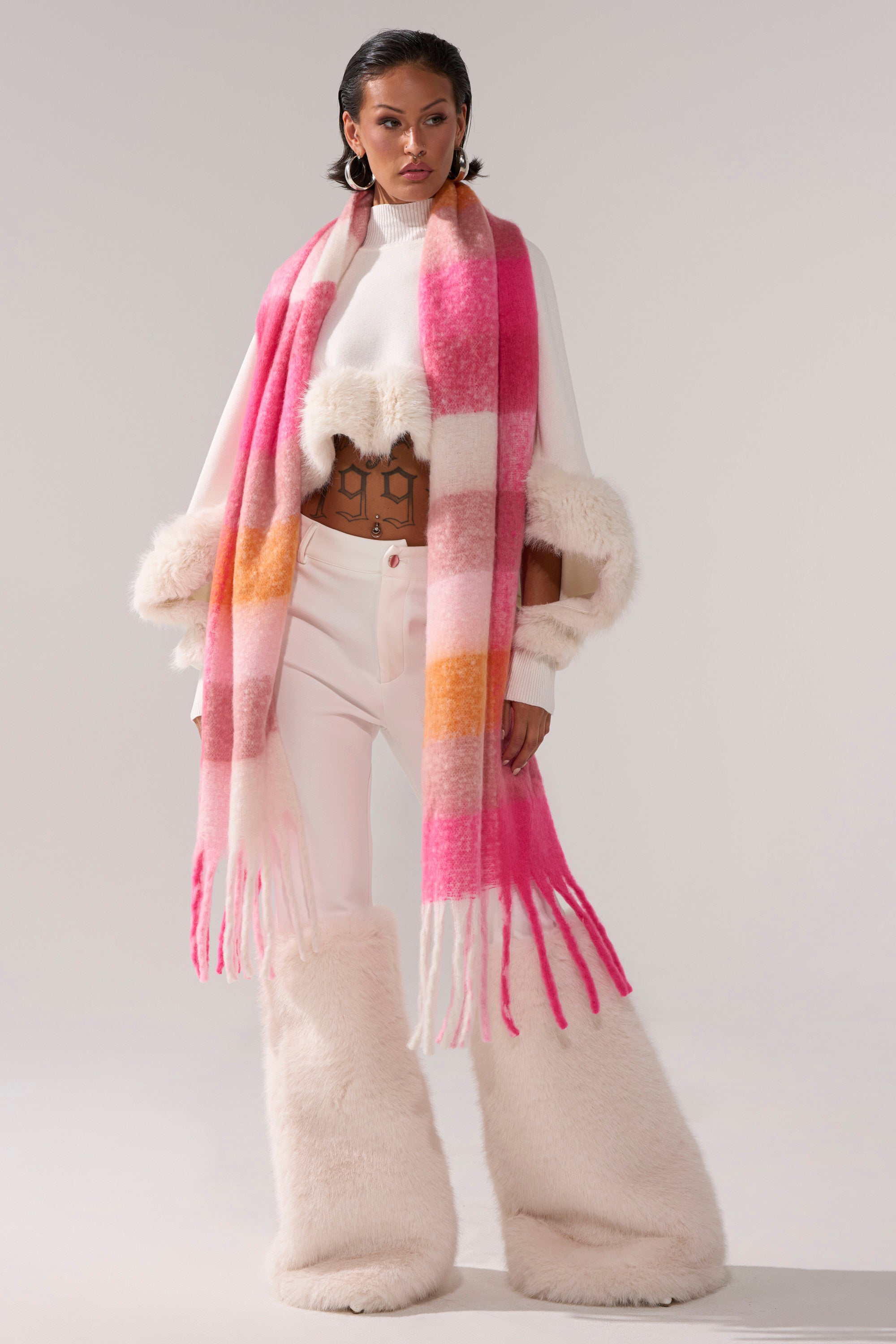 A woman with wavy brown hair and hoop earrings poses in a white cropped sweater with fur trim, high-waisted white pants with fur cuffs, and the IN MILAN BLANKET SCARF featuring a pink, orange, and white plaid pattern.