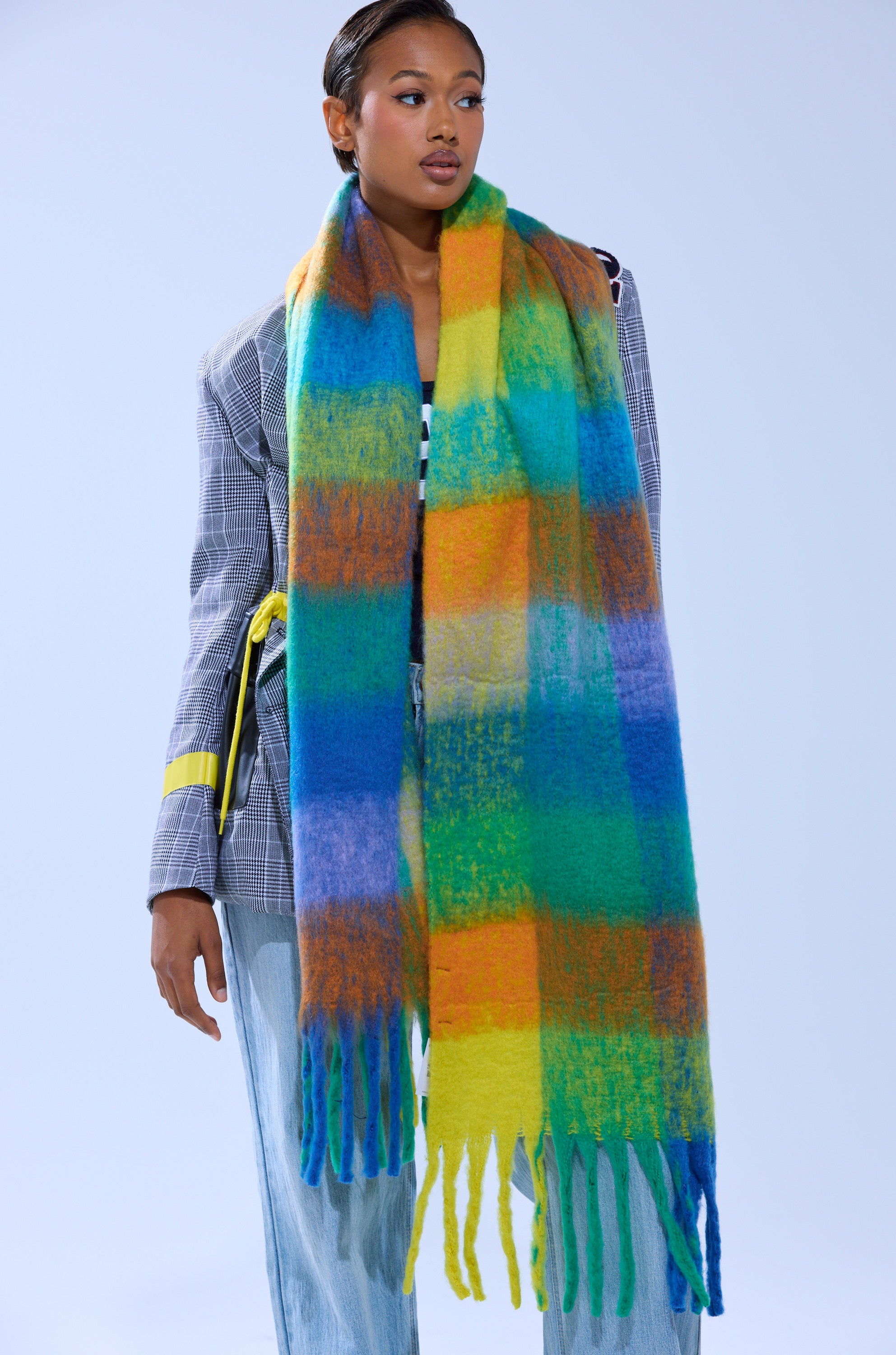 A woman wears the IN PARIS BLANKET SCARF, a large multicolored plaid scarf with fringe in bright yellow, green, blue, and orange, draped over a gray plaid blazer and light blue jeans.