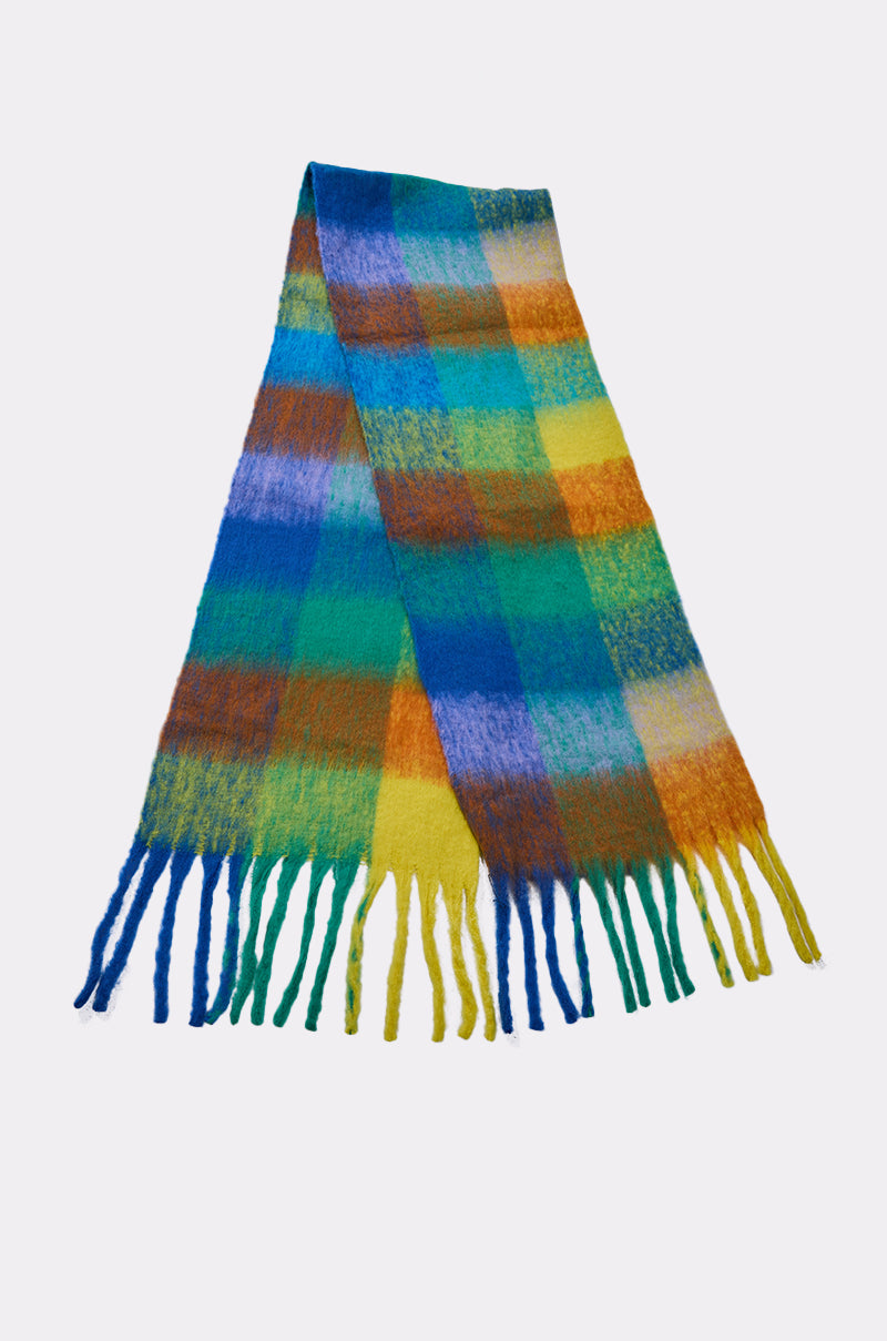 The IN PARIS BLANKET SCARF is a fuzzy scarf with a colorful plaid pattern in blue, green, yellow, and orange hues and fringed ends, shown on a plain white background.