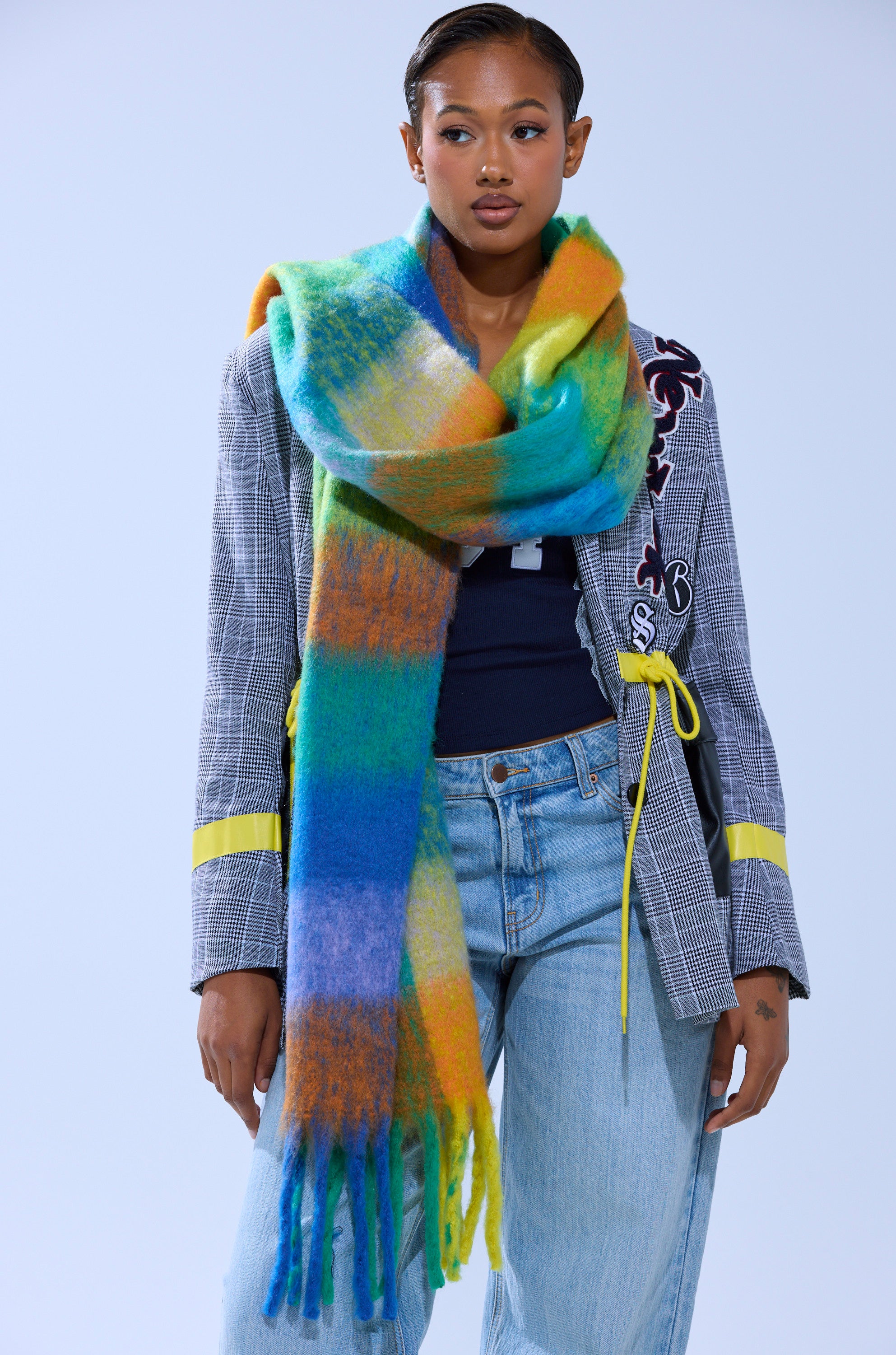 A woman wearing the IN PARIS BLANKET SCARF draped over a checkered blazer with yellow accents, a dark top, and light blue jeans stands against a plain light background.