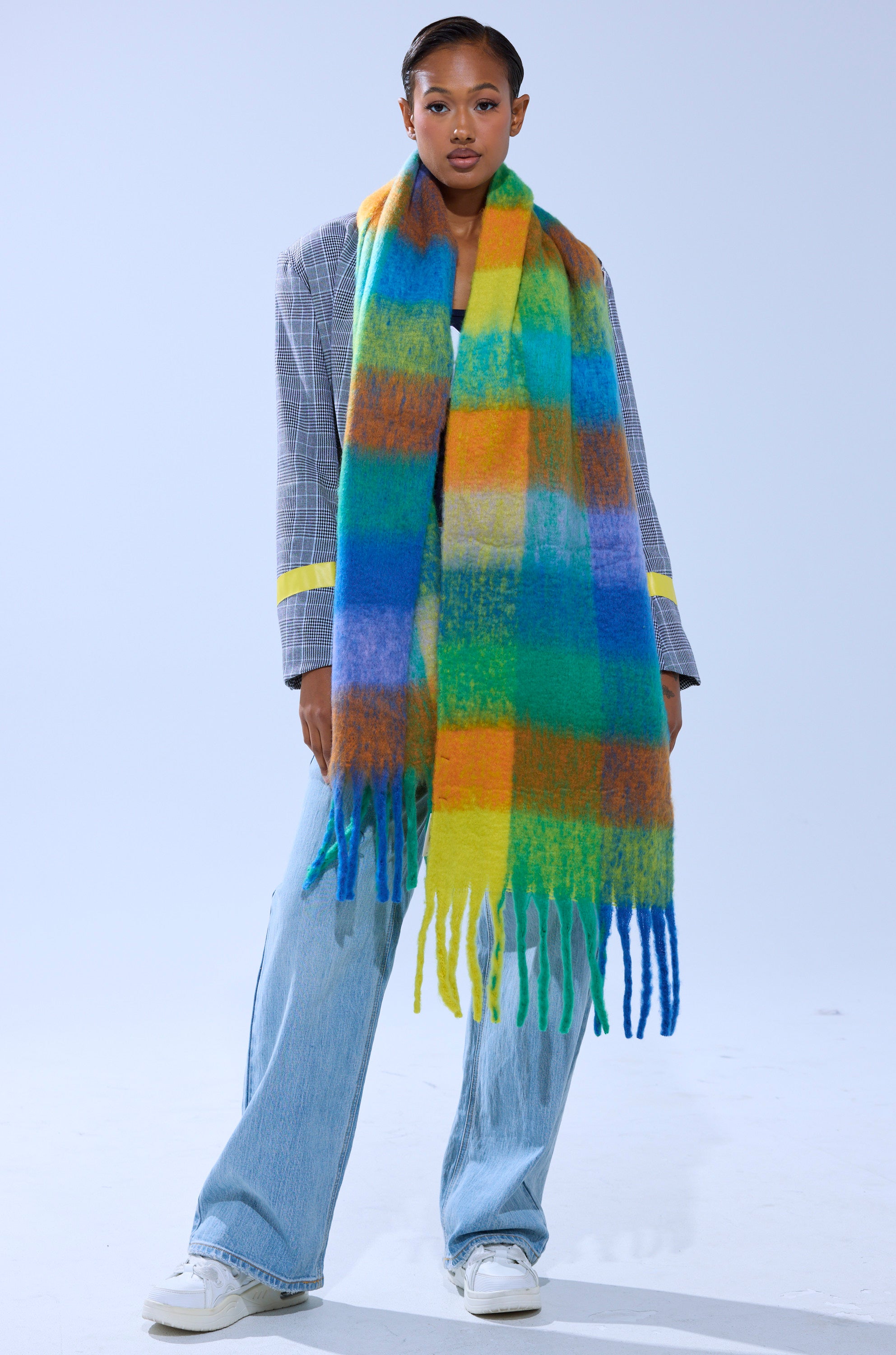 A woman stands against a plain light background, wearing a gray plaid blazer, light blue wide-leg jeans, white sneakers, and the IN PARIS BLANKET SCARF—a large colorful plaid scarf with fringe in green, yellow, orange, and blue.