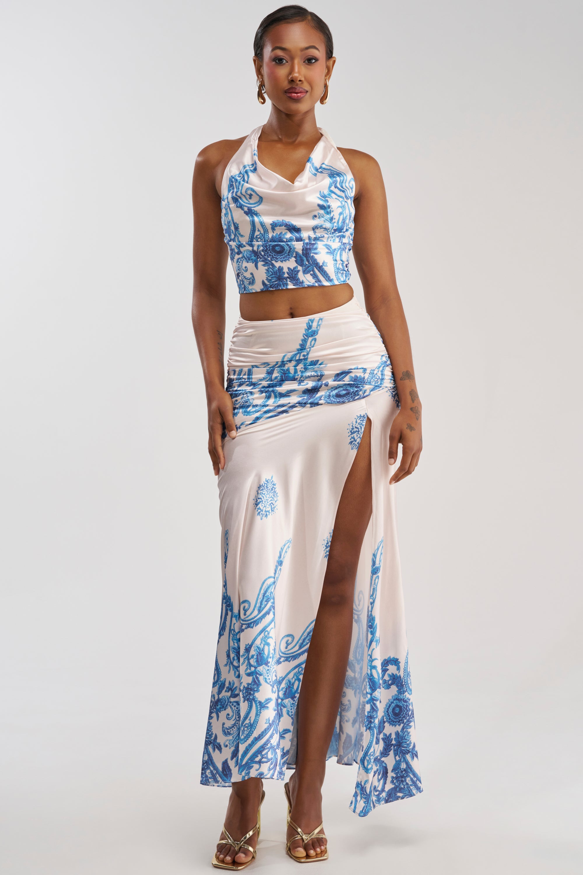 A woman stands against a plain background in gold heels, wearing a sleeveless white crop top paired with the TRINA RUCHED MAXI SKIRT—a matching high-slit summer skirt featuring blue floral patterns.