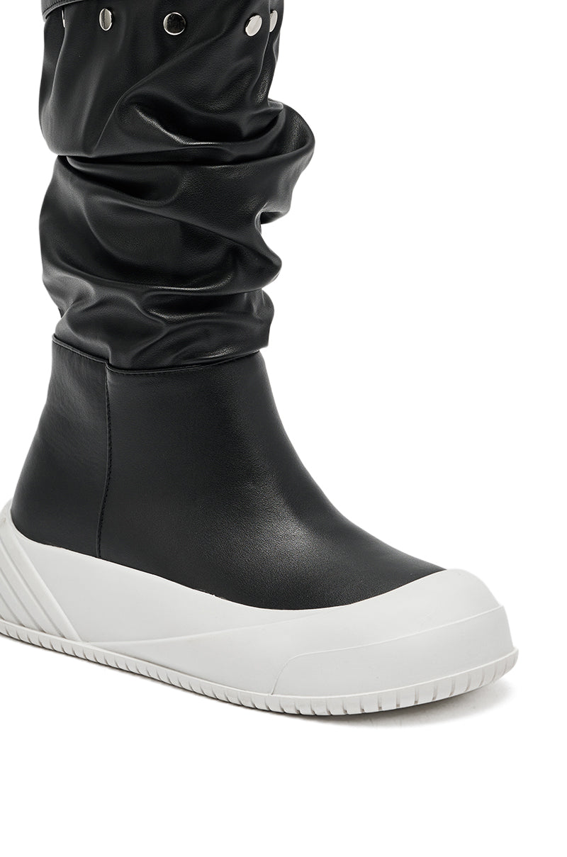A close-up of the AZALEA WANG SMARTER BLACK RUCHED PU FLAT BOOT, a knee-high black slouchy style with a thick white rubber sole and toe cap, plus metal eyelets along the top.