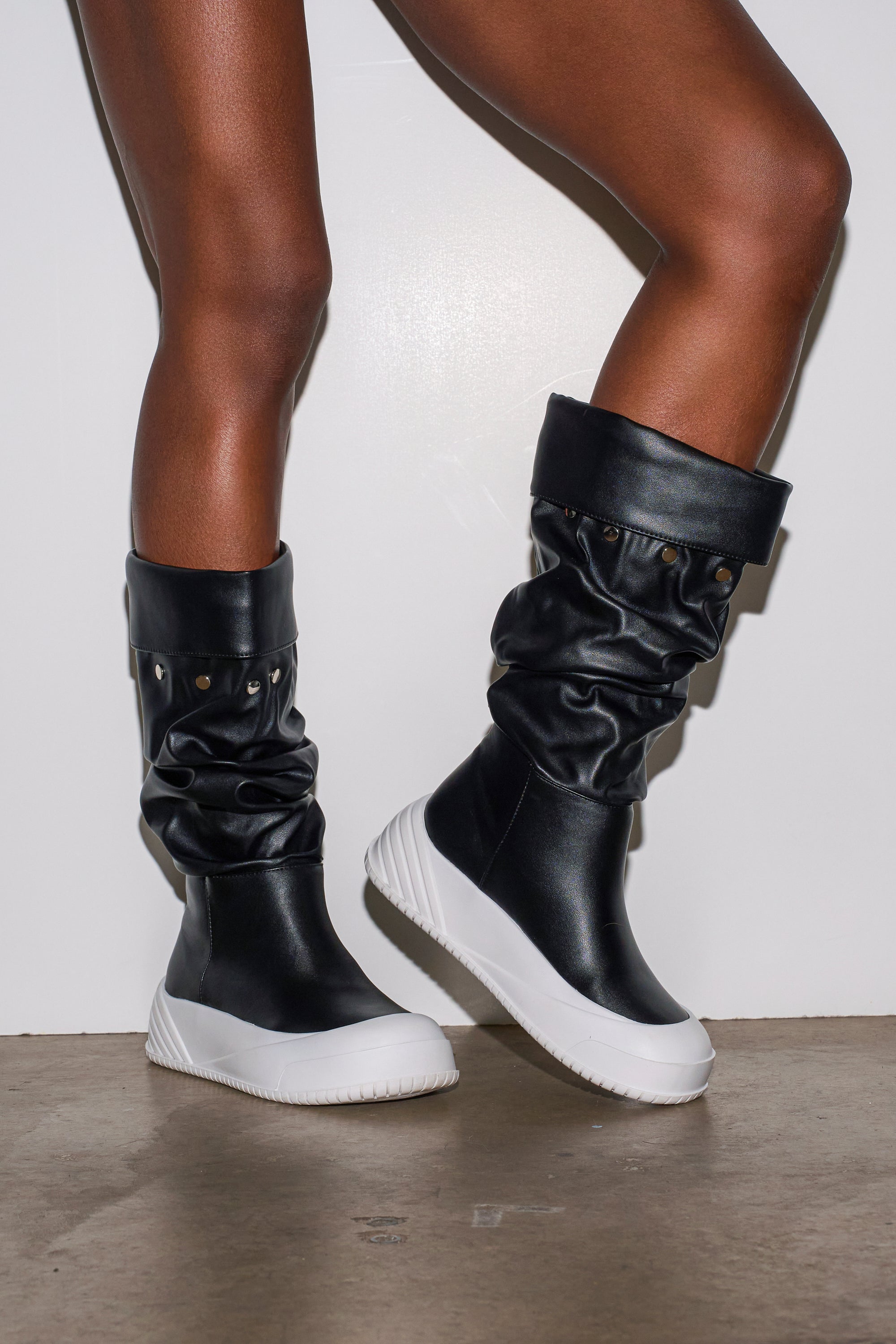 A person stands on a concrete floor wearing the AZALEA WANG SMARTER BLACK RUCHED PU FLAT BOOT, featuring white chunky soles and metal stud details, shown from mid-thigh down against a plain white background.
