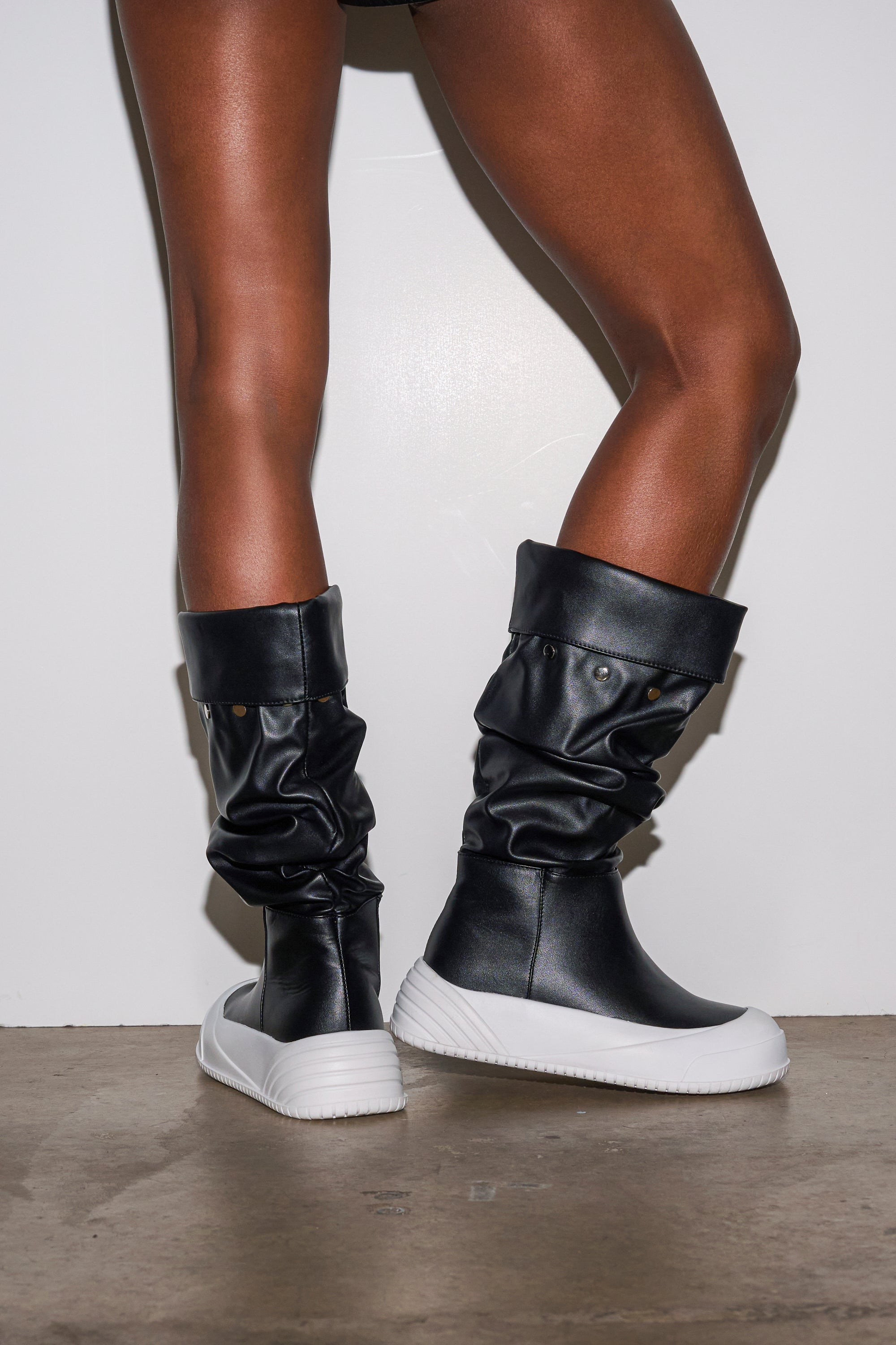 A person stands on a concrete floor, wearing the AZALEA WANG SMARTER BLACK RUCHED PU FLAT BOOT with thick white soles, set against a plain white background.
