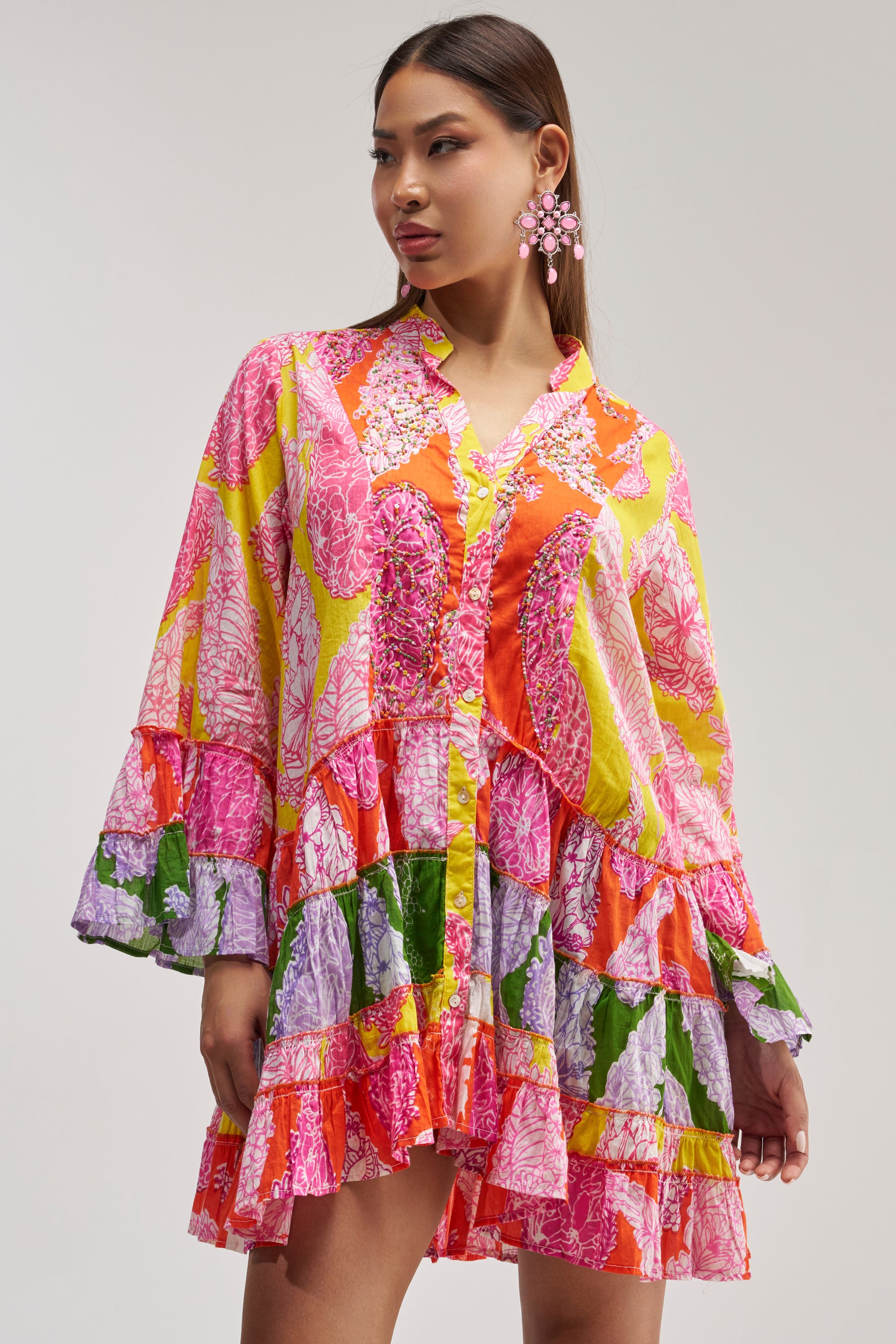 A woman with long hair and bold earrings stands against a plain background, wearing the DANCING TILL SUNRISE FLORAL LONG SLEEVE DRESS—a vibrant festival dress featuring colorful pink, red, yellow, green, and purple floral patterns.