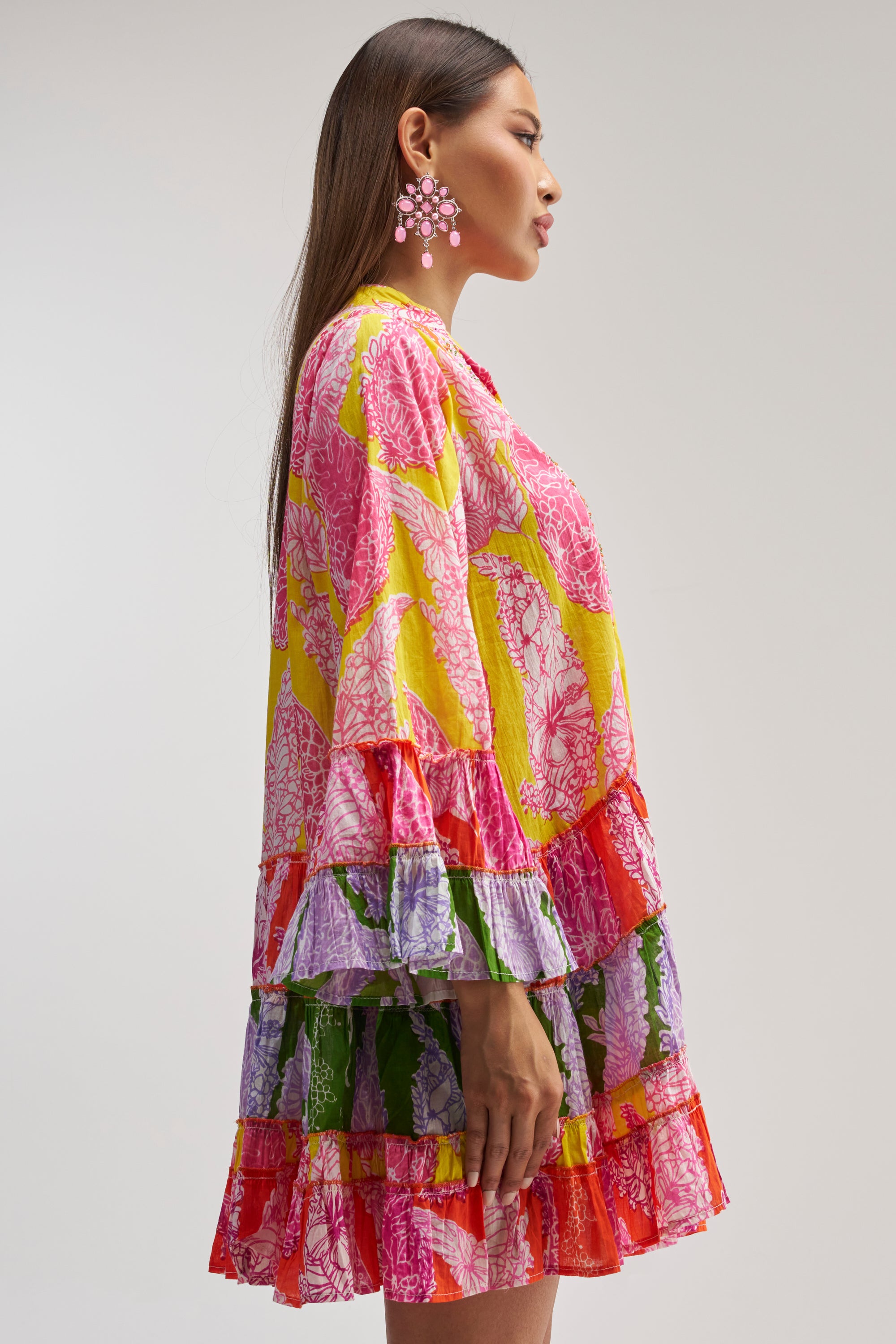 A woman in profile models the DANCING TILL SUNRISE FLORAL LONG SLEEVE DRESS, featuring a vibrant floral pattern with ruffled sleeves and skirt. She has long, straight brown hair and large pink flower earrings, standing against a plain light background.