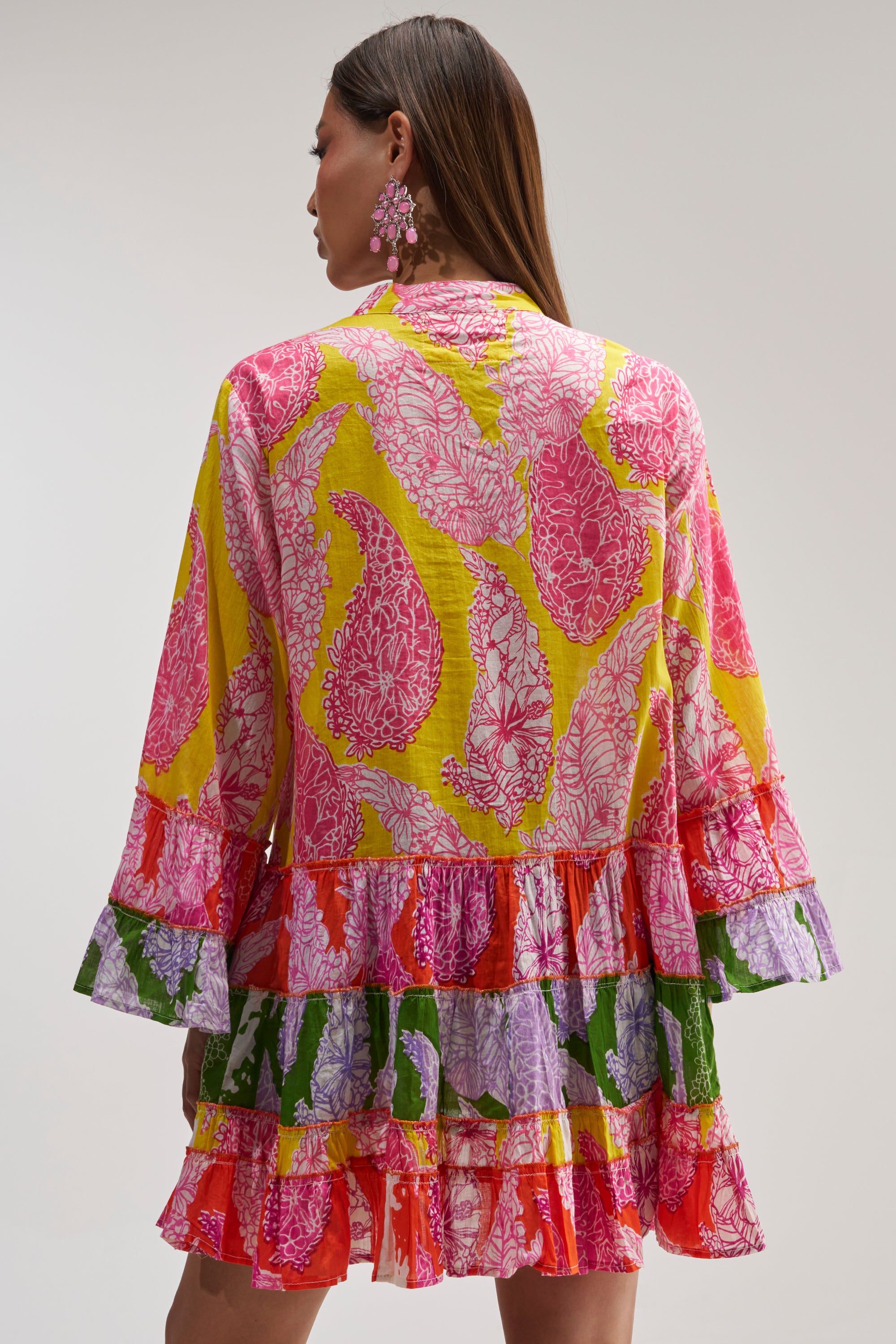 A woman facing away from the camera wears the DANCING TILL SUNRISE FLORAL LONG SLEEVE DRESS, featuring colorful floral prints, ruffled layers, and pairs it with pink statement earrings.